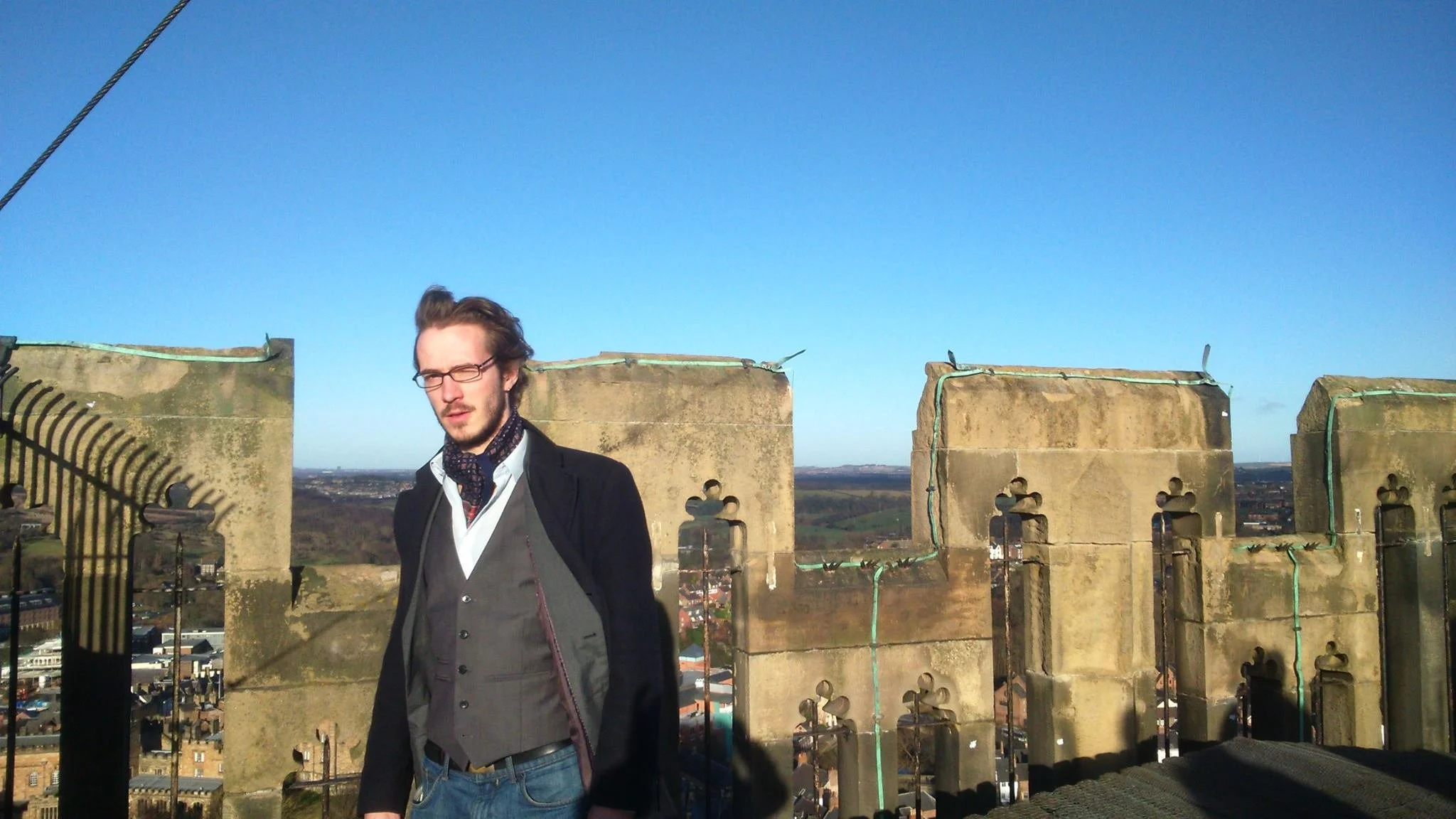 A man with glasses and long hair standing on a rooftop or high balcony with stone crenellations, overlooking a cityscape under a clear blue sky.