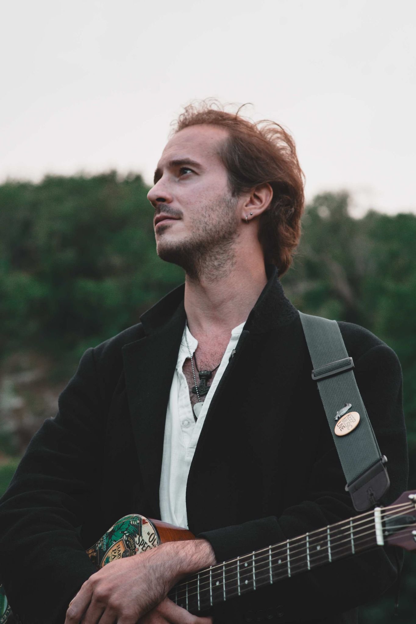 A man with reddish-brown hair and a slight beard, wearing a dark jacket over a white shirt, holding an acoustic guitar outdoors with a blurred green background.
