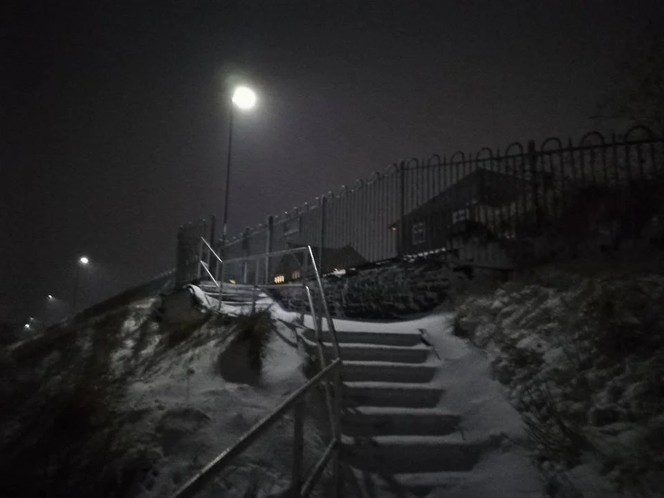 Snow-covered outdoor staircase at night with a metal railing, overlooking a foggy landscape with lights, barbed wire, and a house in the distance under a bright full moon.