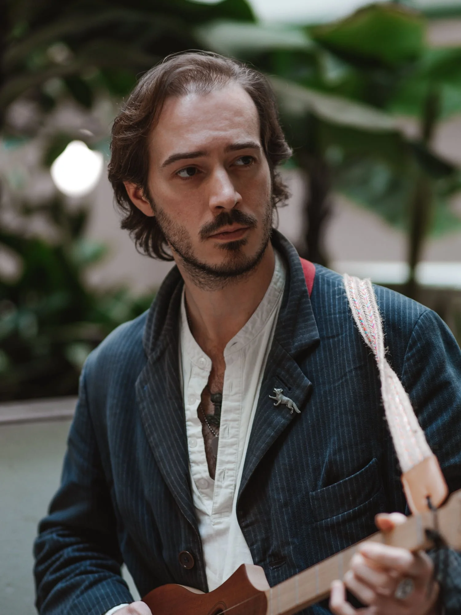A man with shoulder-length wavy brown hair, facial hair, and light skin, plays a ukulele outdoors with green foliage in the background.