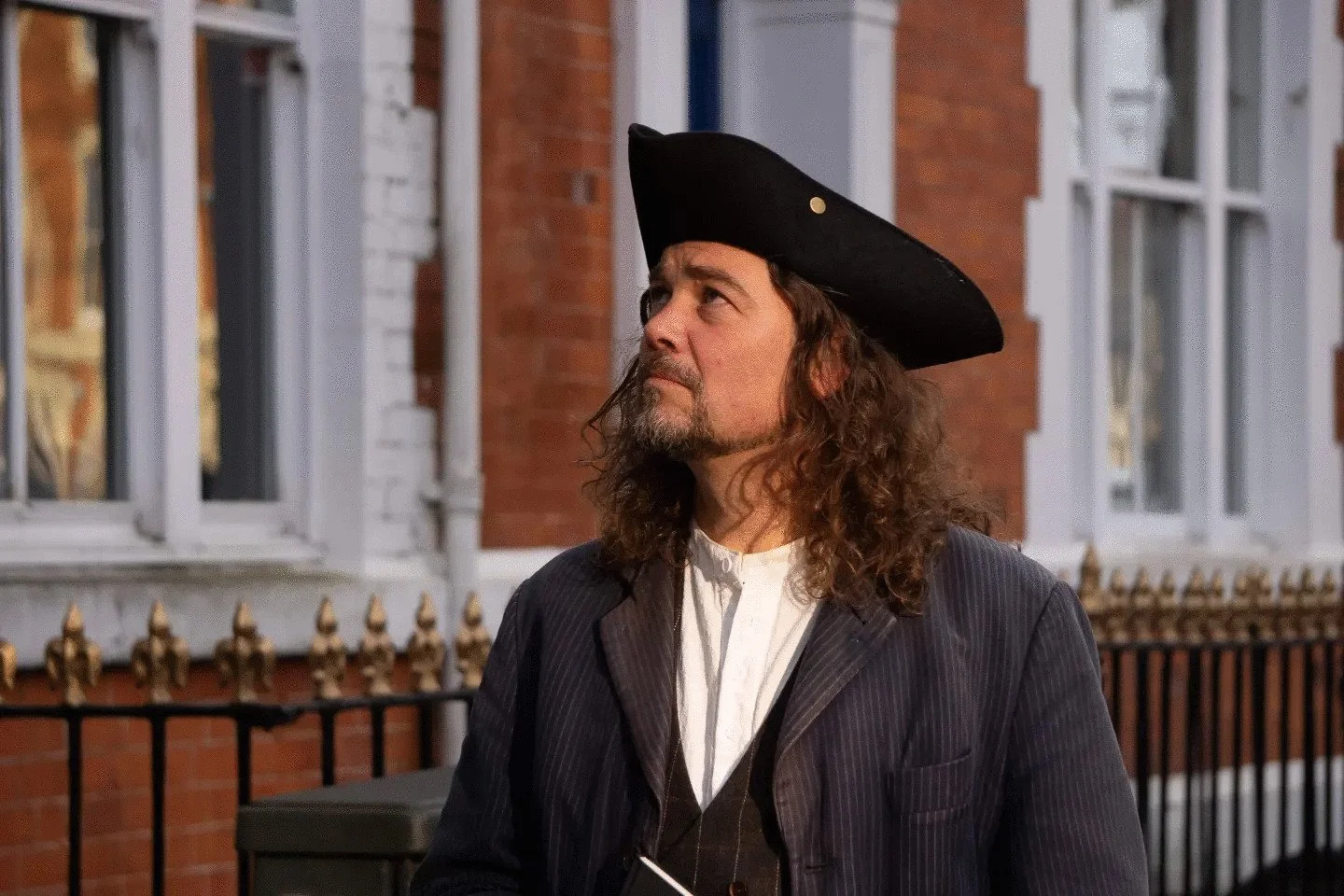 A man dressed as a pirate with a tricorn hat, long curly hair, a beard, and period costume, standing outdoors in front of a brick and white windowed building.