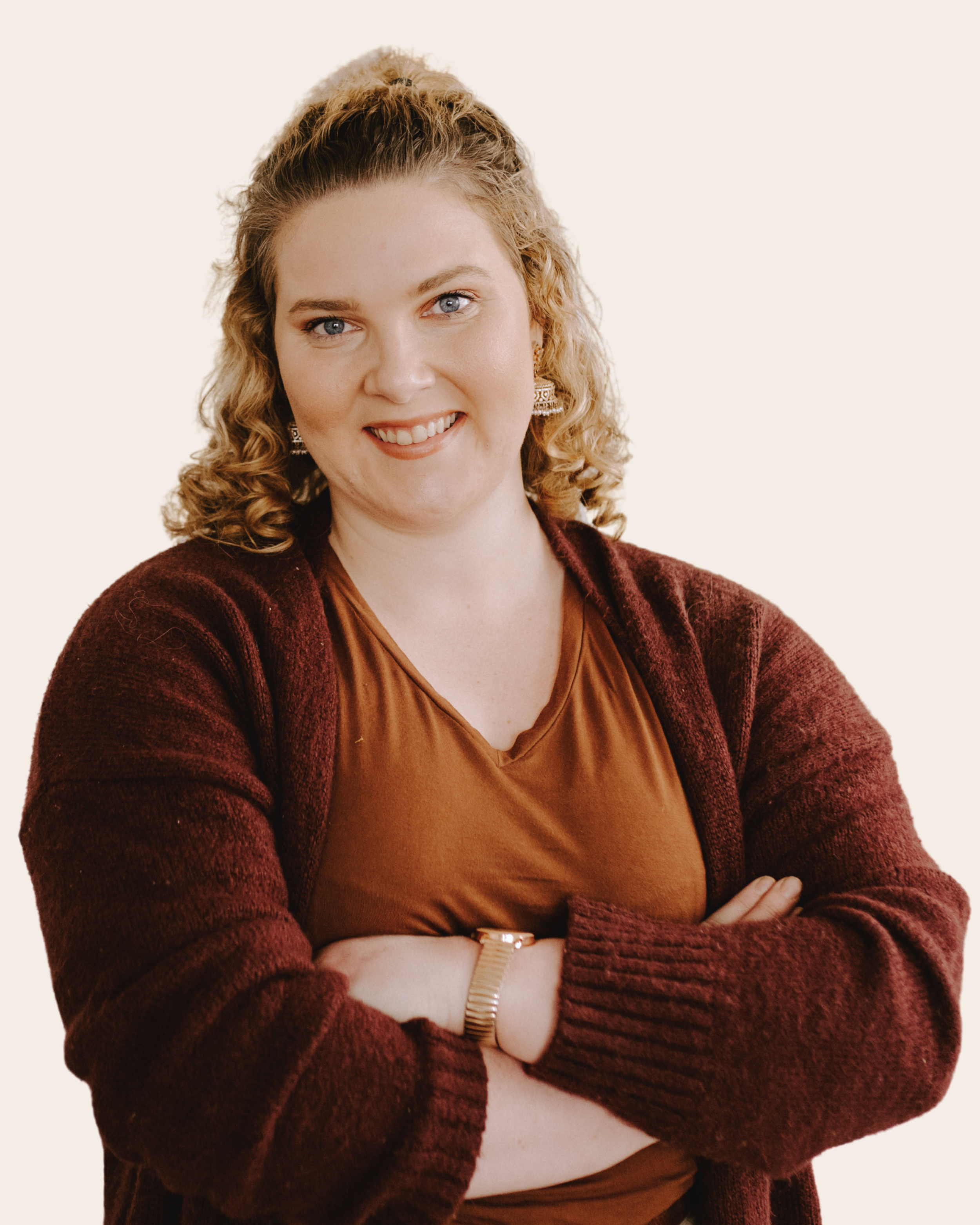 A woman with curly blonde hair, blue eyes, and fair skin, smiling with arms crossed, wearing a rust-colored top, a maroon cardigan, earrings, and a gold watch, standing against a beige background.