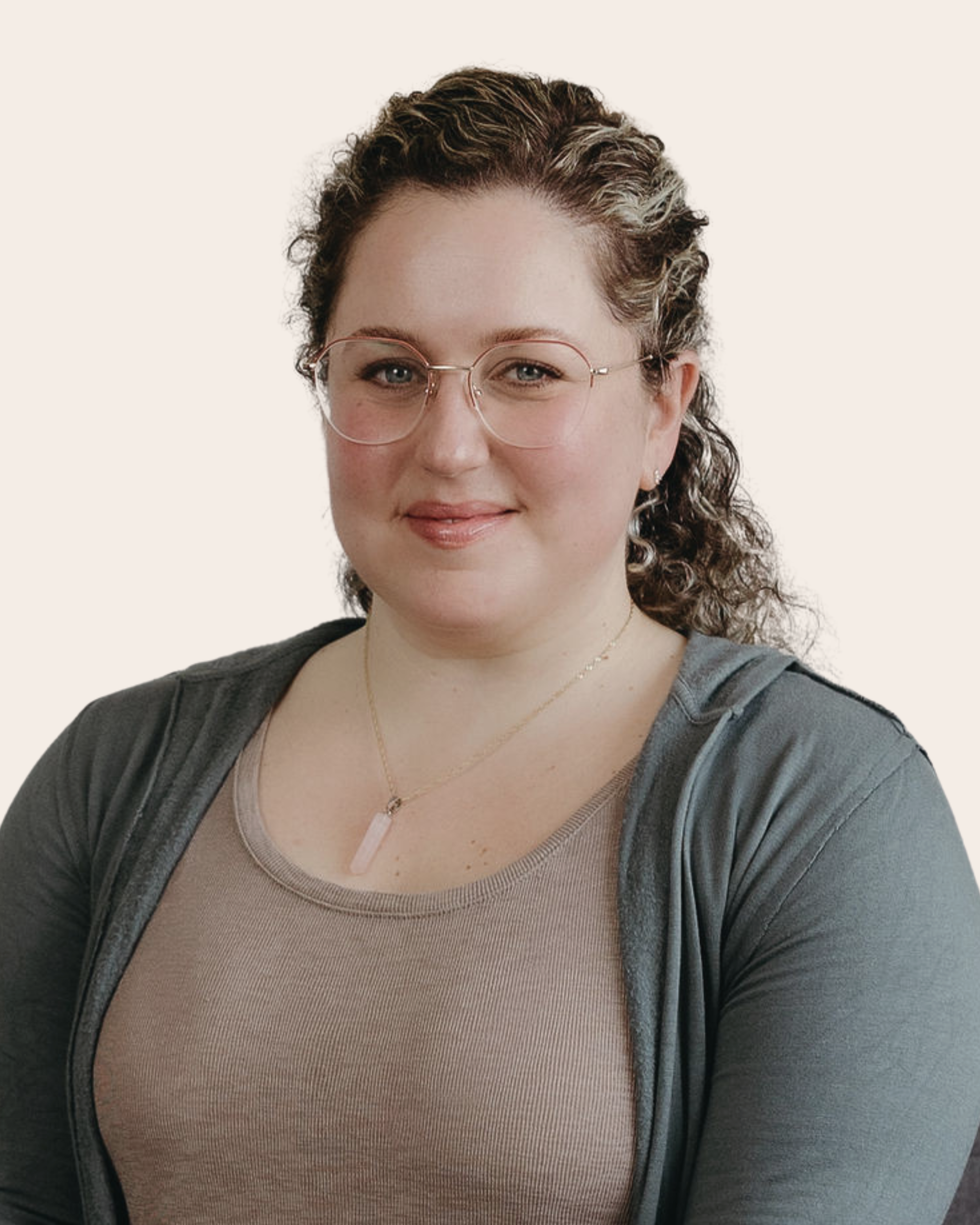 A woman with curly hair, wearing glasses with round frames, a beige top, and a gray jacket, smiling at the camera.