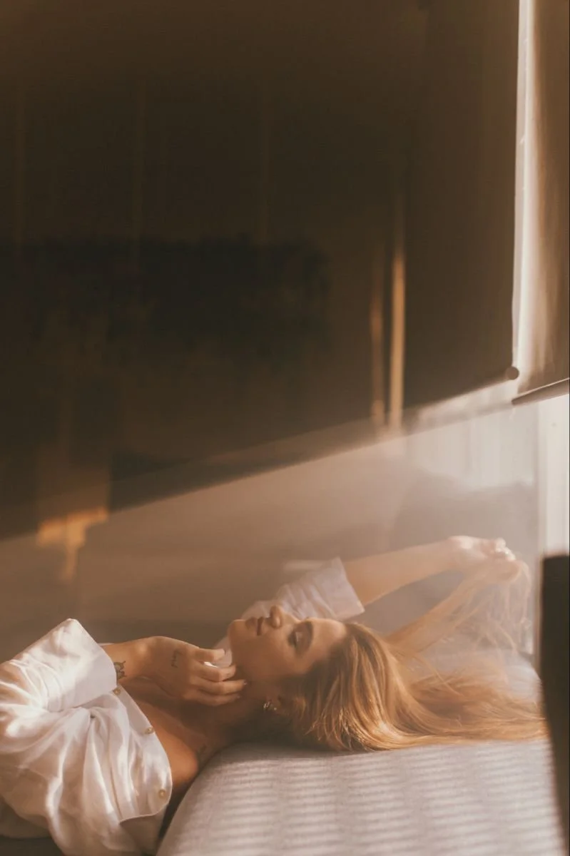 A woman with long reddish hair lying on a bed with her hand touching her face, wearing a white shirt, in a warm-toned room with sunlight filtering in.