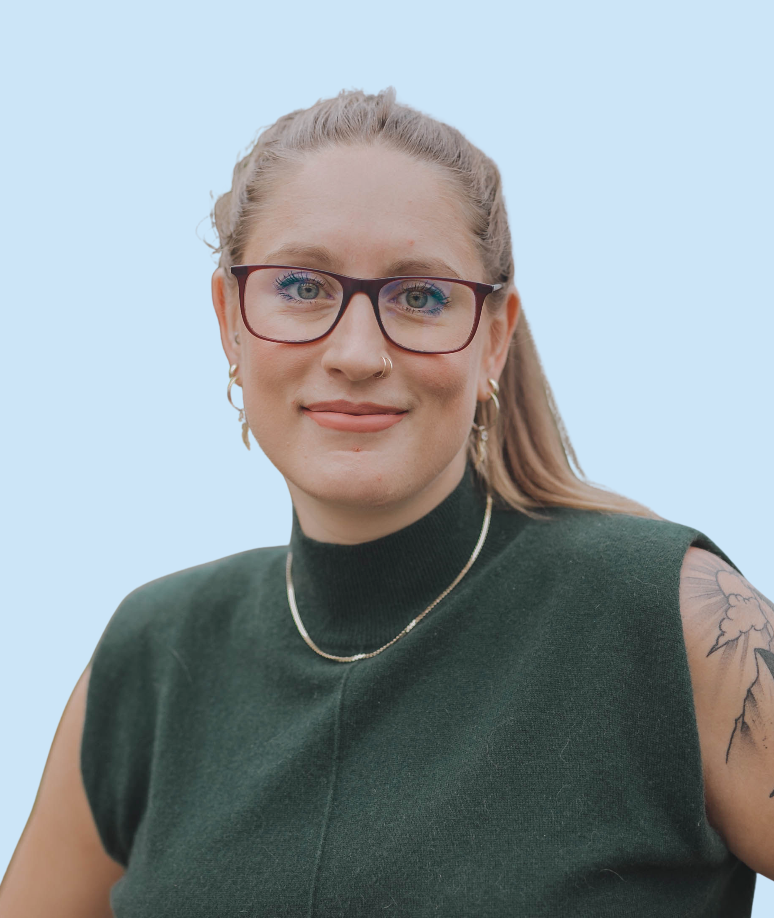 A woman with glasses, nose piercing, earrings, necklace, and tattoo on her shoulder, wearing a dark sleeveless top, smiling against a light background.