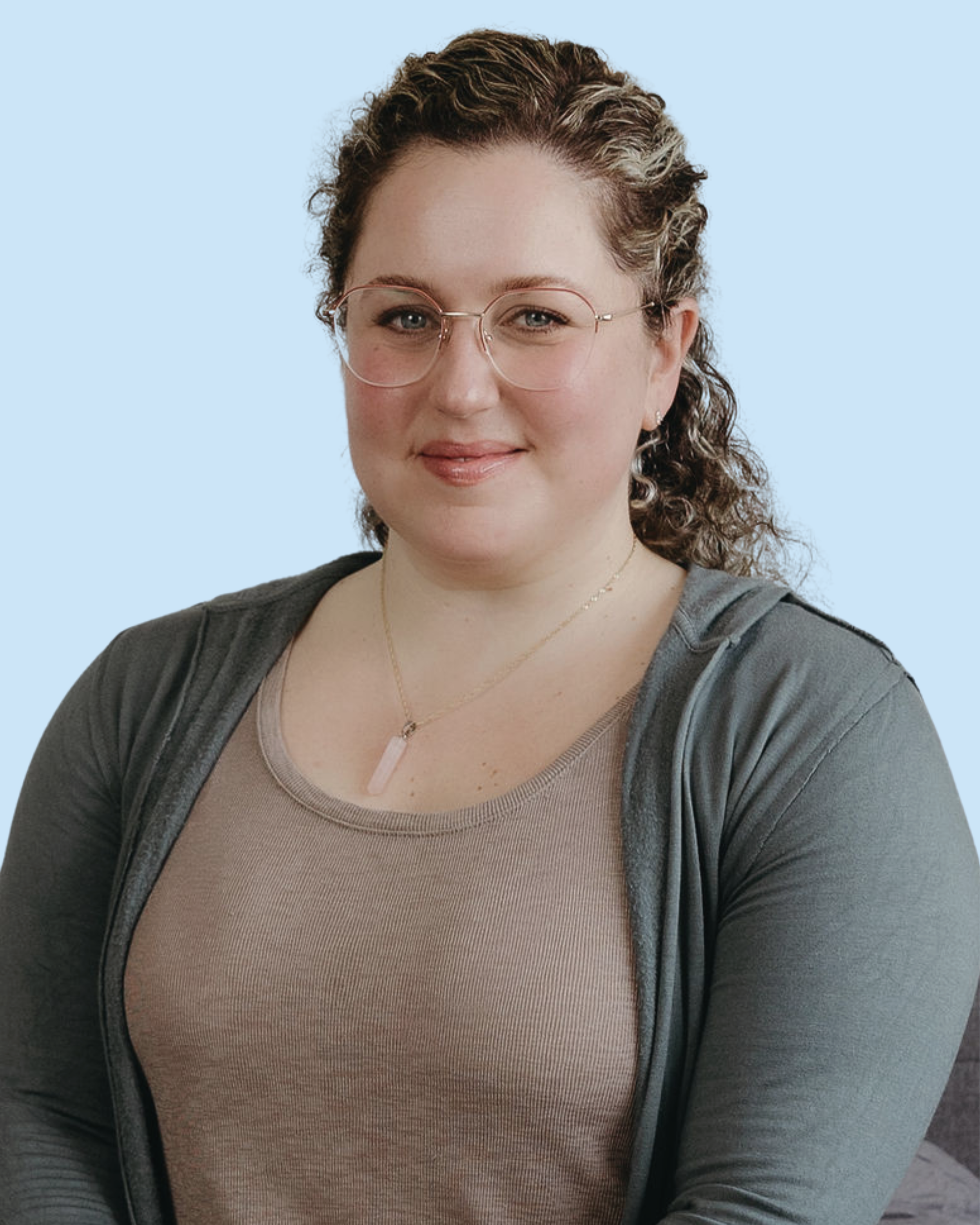 A woman with curly brown hair, glasses, and light makeup, smiling, wearing a beige top, a dark gray cardigan, and a pink quartz necklace, against a light blue background.