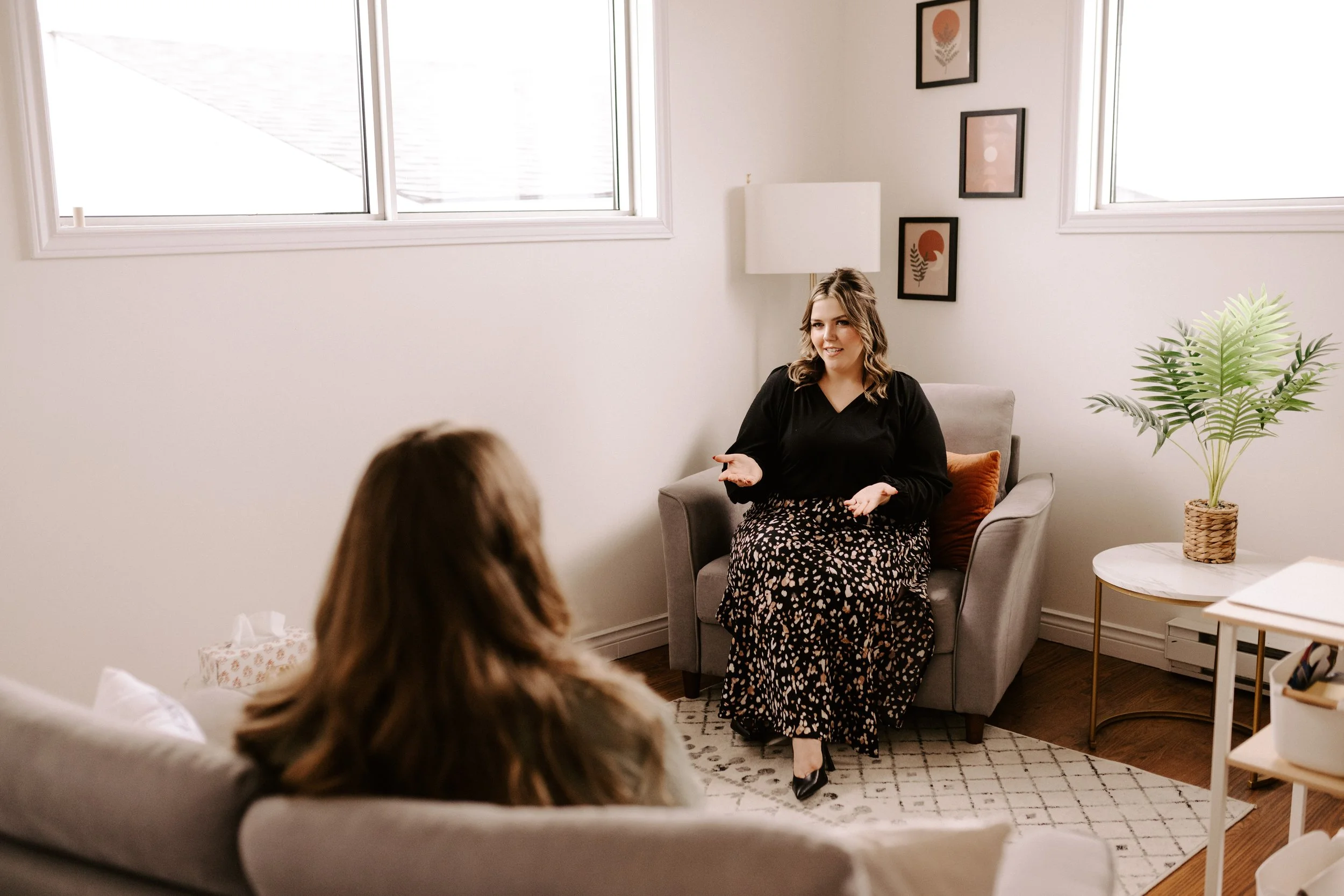 Two women having a conversation in a living room, one sitting on a couch with her back partially turned, the other sitting on an armchair, gesturing with her hands.