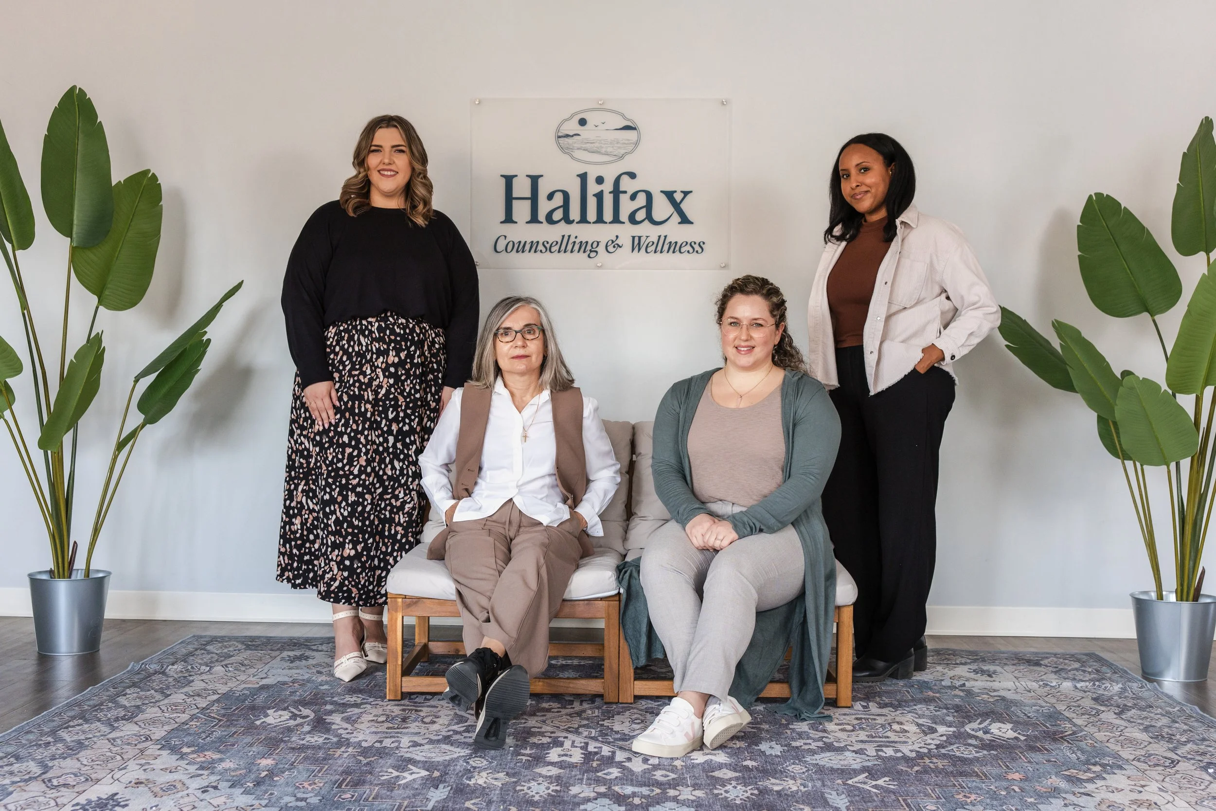 Group of five women in an office setting, sitting and standing in front of a sign that says 'Halifax Counseling & Wellness,' with potted plants on either side.