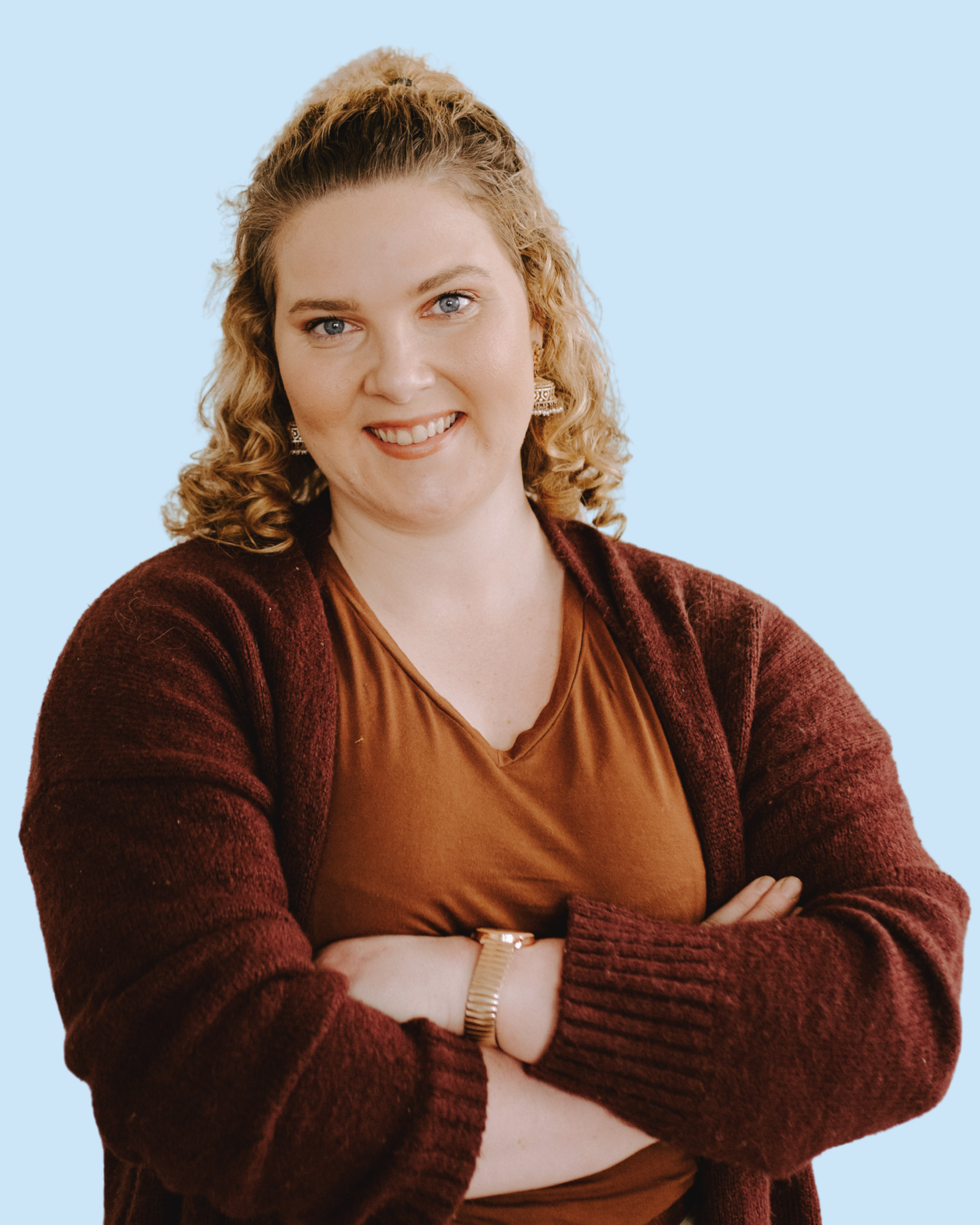 A woman with curly blonde hair, blue eyes, and fair skin, smiling and crossing her arms, wearing a brown V-neck shirt, maroon cardigan, earrings, and a watch, against a light blue background.