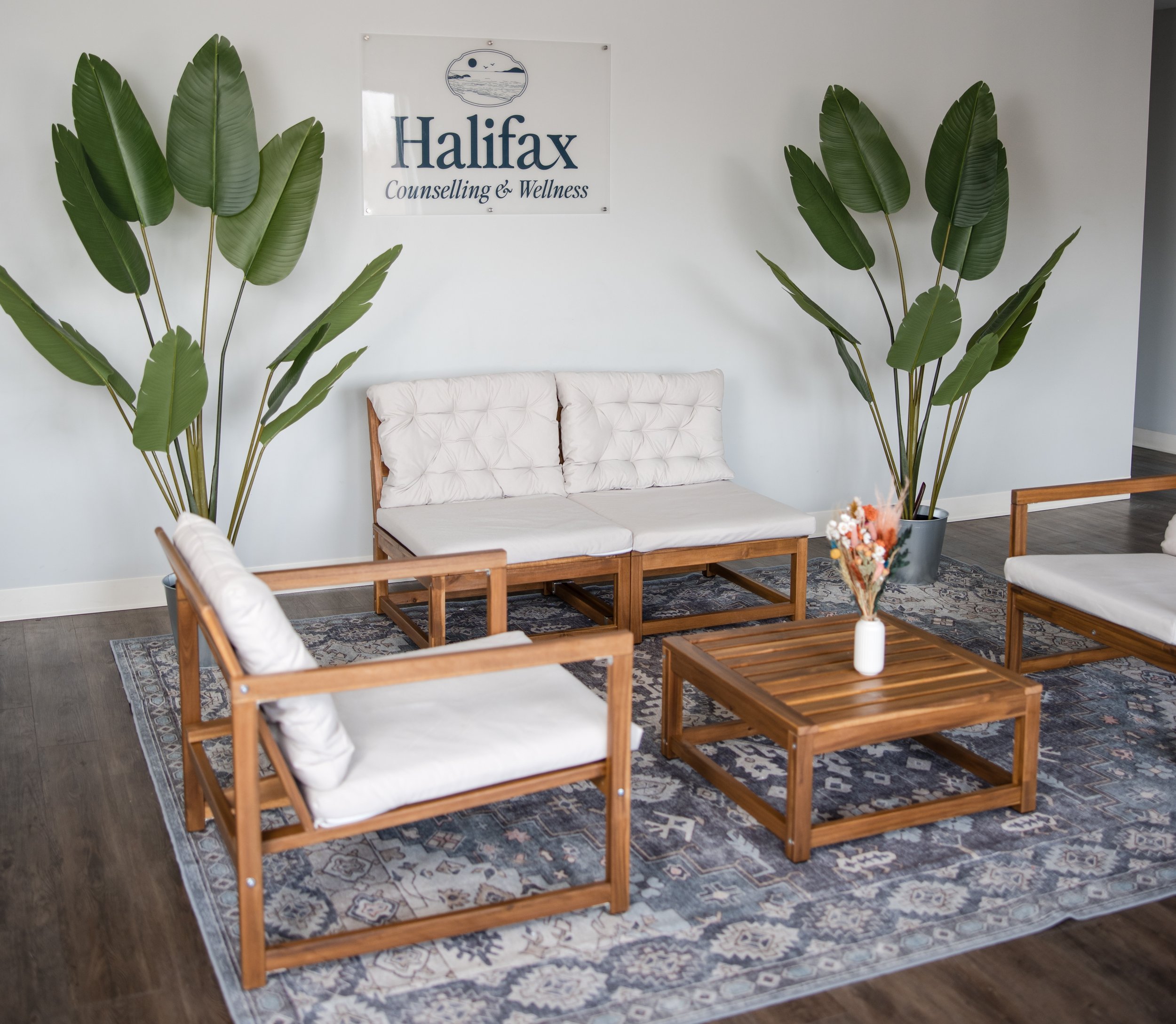Waiting room with wooden furniture, white cushions, large potted tropical plants, and a Halifax Counselling & Wellness sign on the wall.