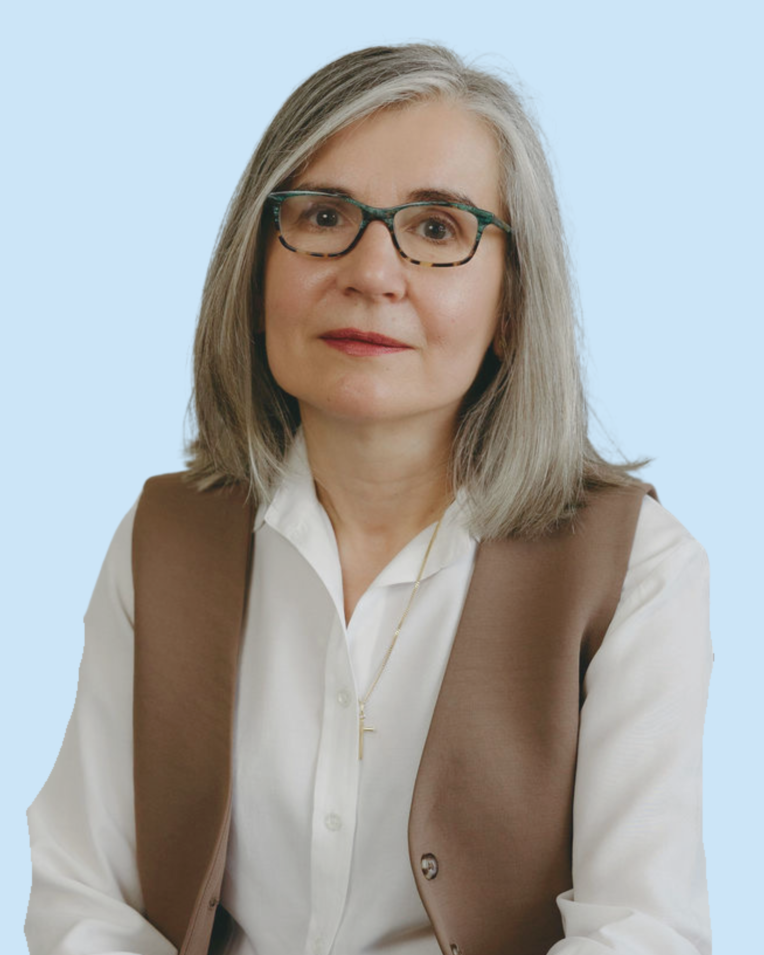 A middle-aged woman with gray shoulder-length hair, glasses, wearing a white blouse and brown vest, posing against a light blue background.