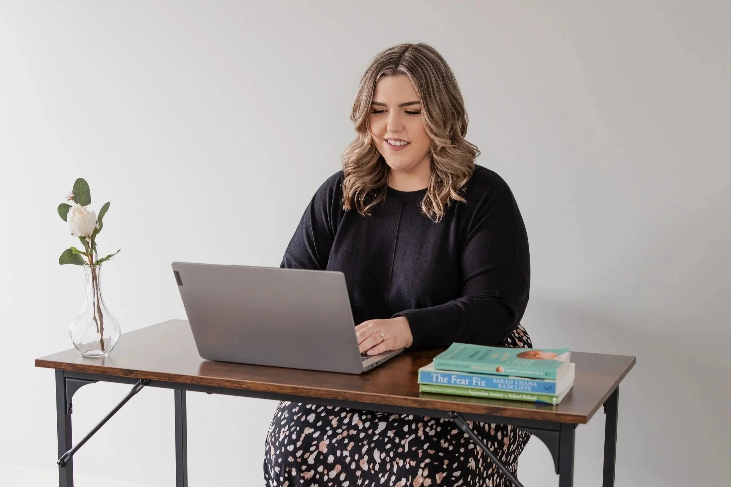 A woman sitting at a desk working on a laptop, with a vase of white flowers and several books on the desk.