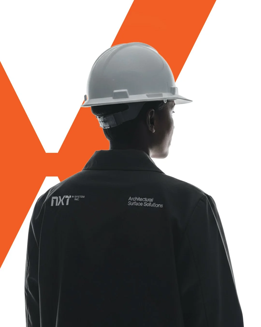 A person wearing a white construction helmet and black jacket with the text 'NXT Architecture Surface Solutions' on the back, standing with their back to the camera and looking to the right.