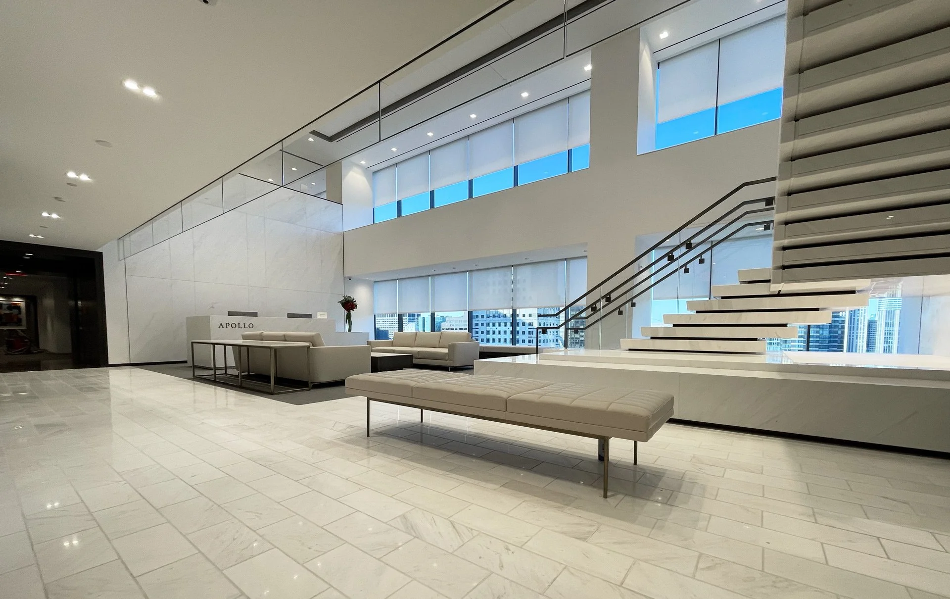 Modern lobby with white marble floors and walls, large windows with city views, white sofas, a white bench, and a staircase with horizontal slats.