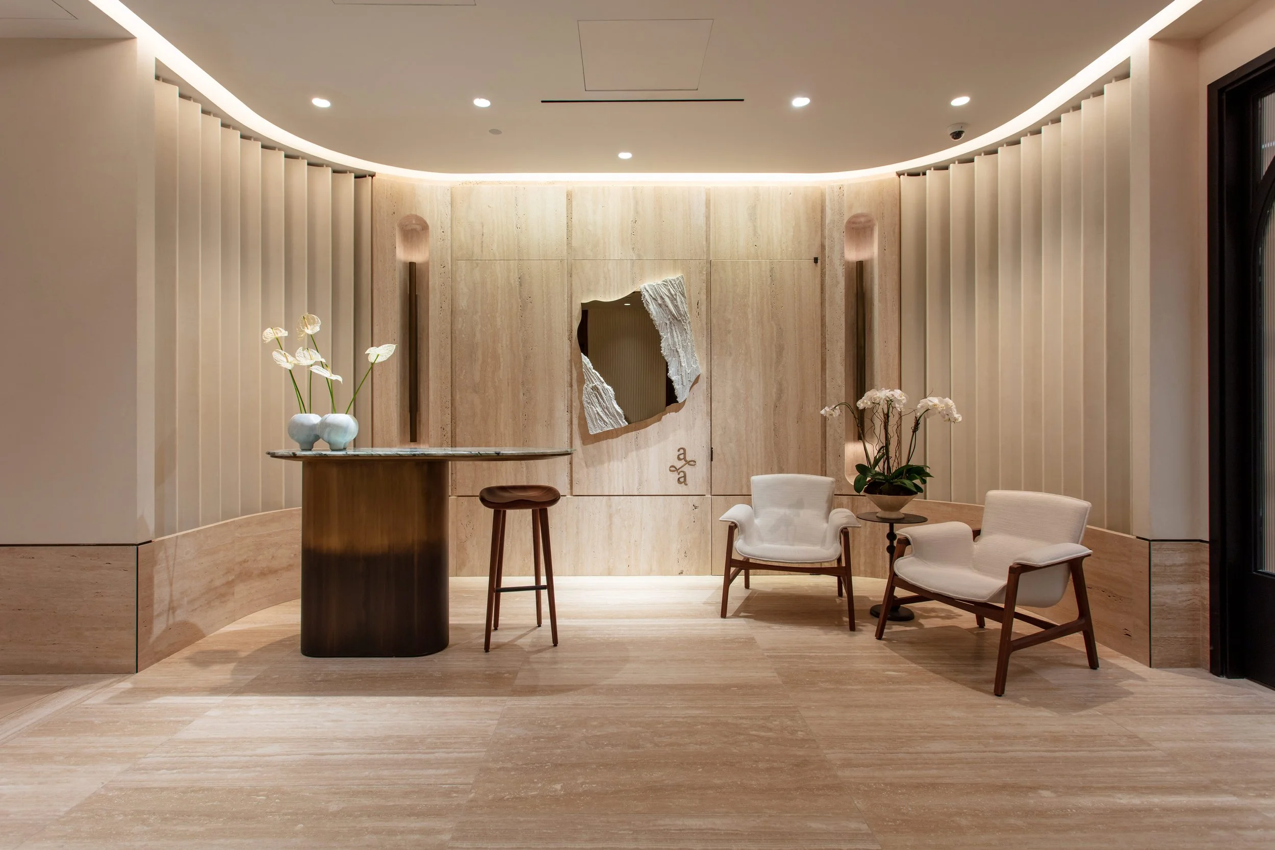 Minimalist hotel lobby with beige marble walls and floor, two white armchairs, a small round table with orchids, a curved reception desk, and a decorative mirror on the wall.