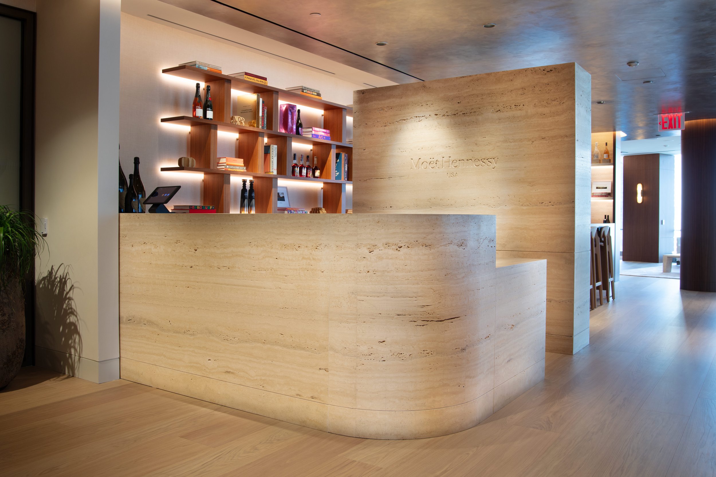 A modern hotel reception area with a curved beige stone counter, a wooden shelf with books and bottles of alcohol, and soft lighting, with a Moët Hennessy sign on the wall.