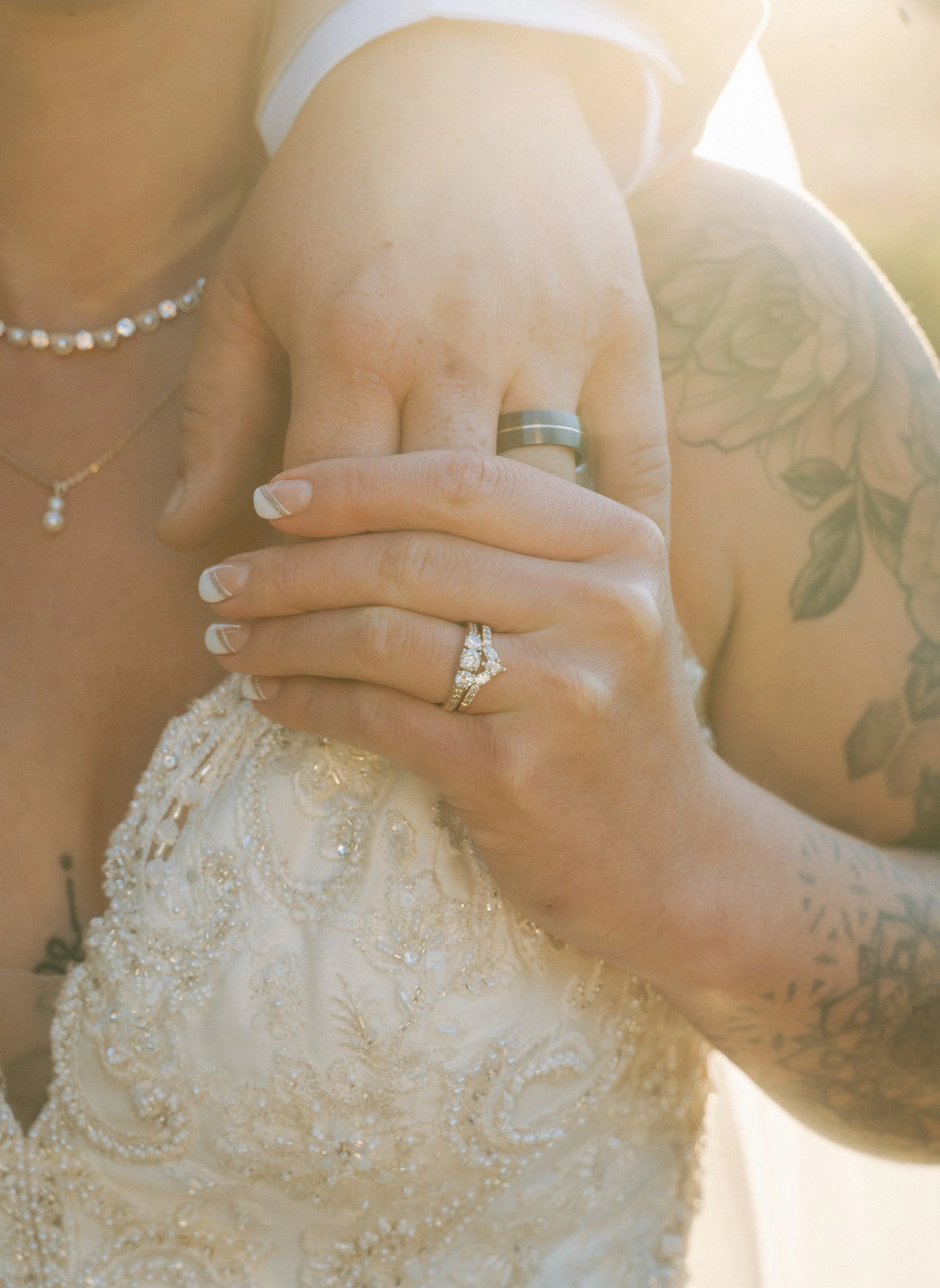 Close-up of a woman embracing herself, showing her wedding and engagement rings, with her nails painted French tips, wearing pearl jewelry and a beaded wedding dress. Part of her arm with a floral tattoo is visible.