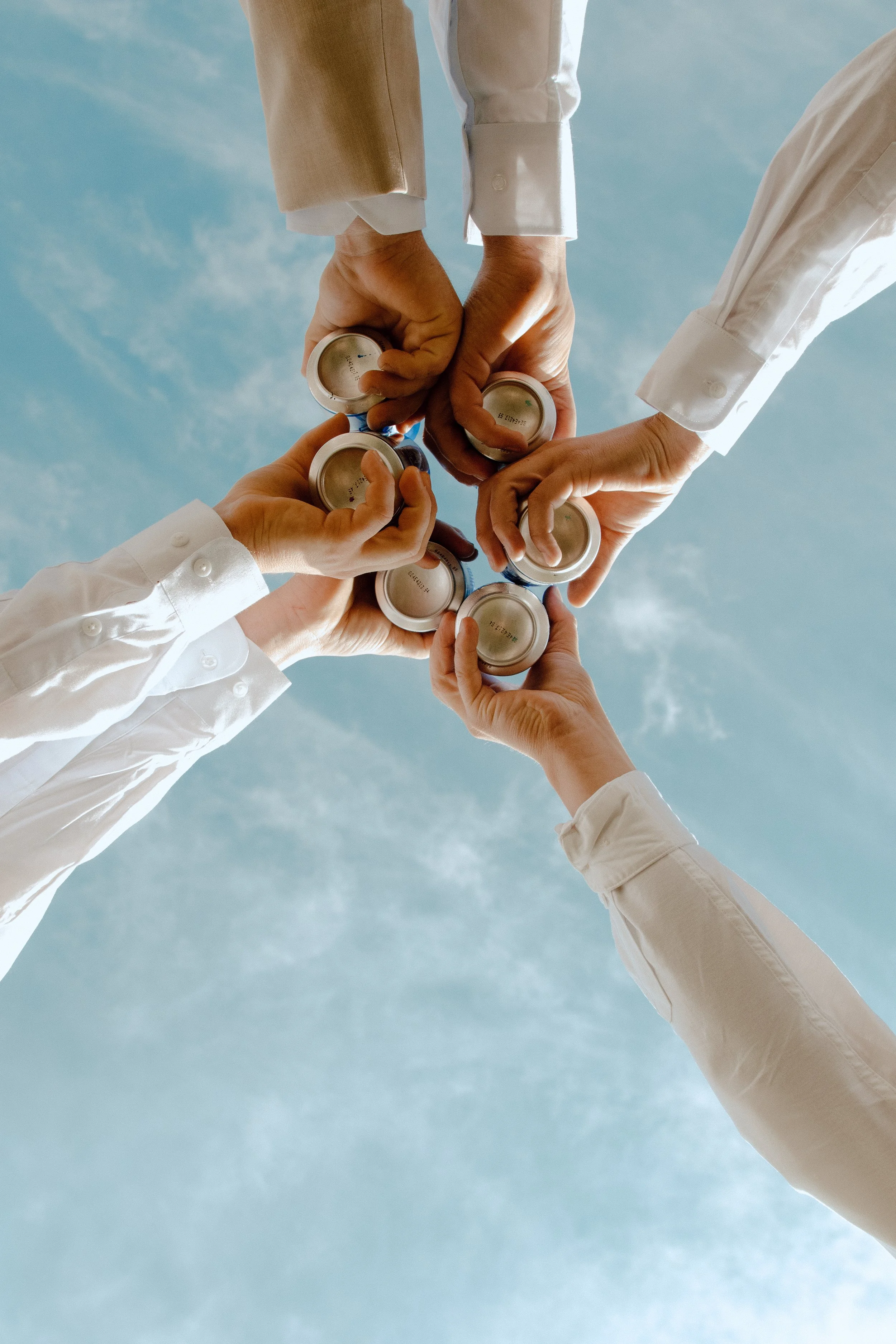 Multiple hands holding small cans, forming a circle against a blue sky with clouds.