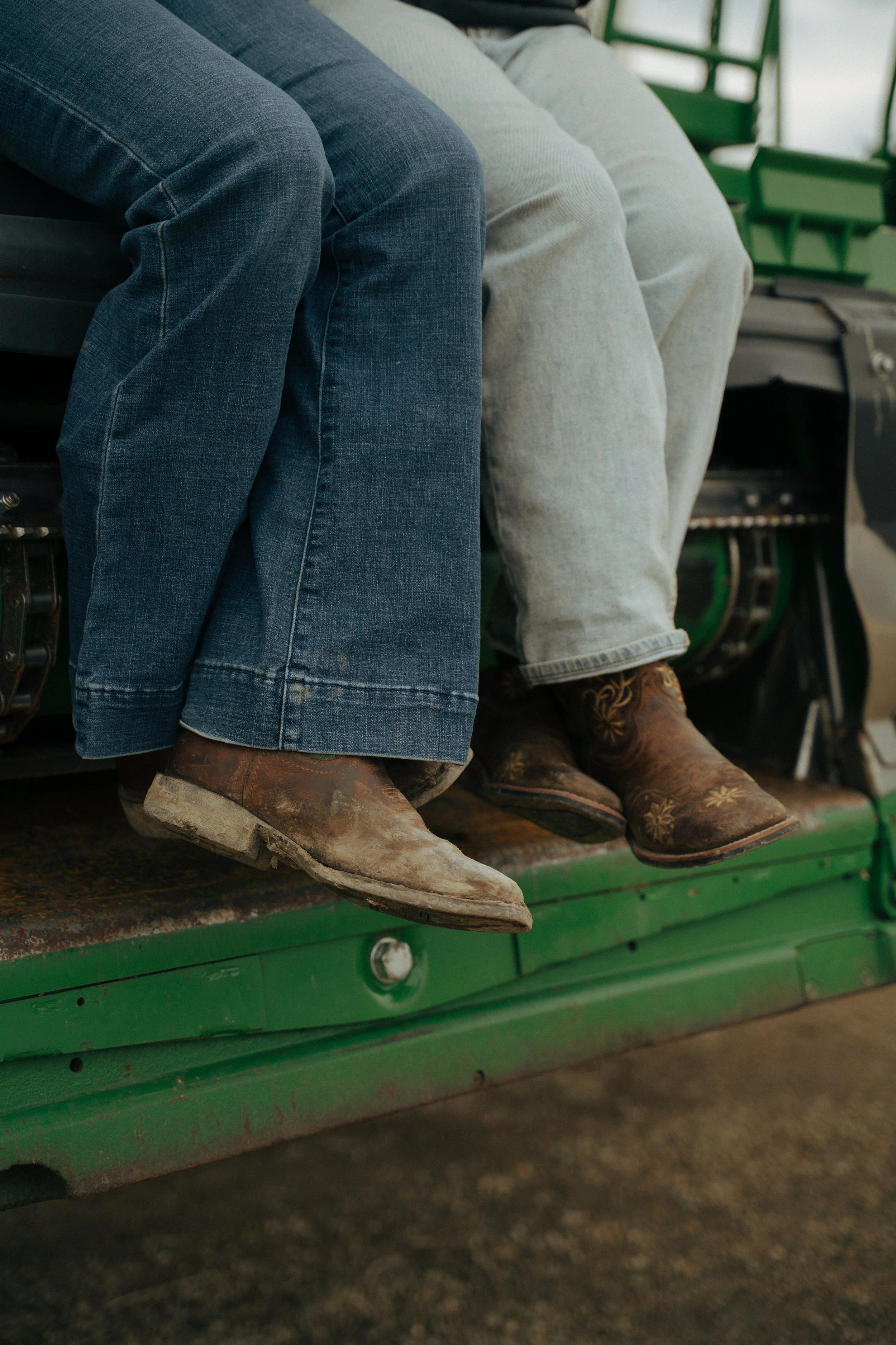 People sitting on a green farm machinery, focusing on their legs and worn cowboy boots.