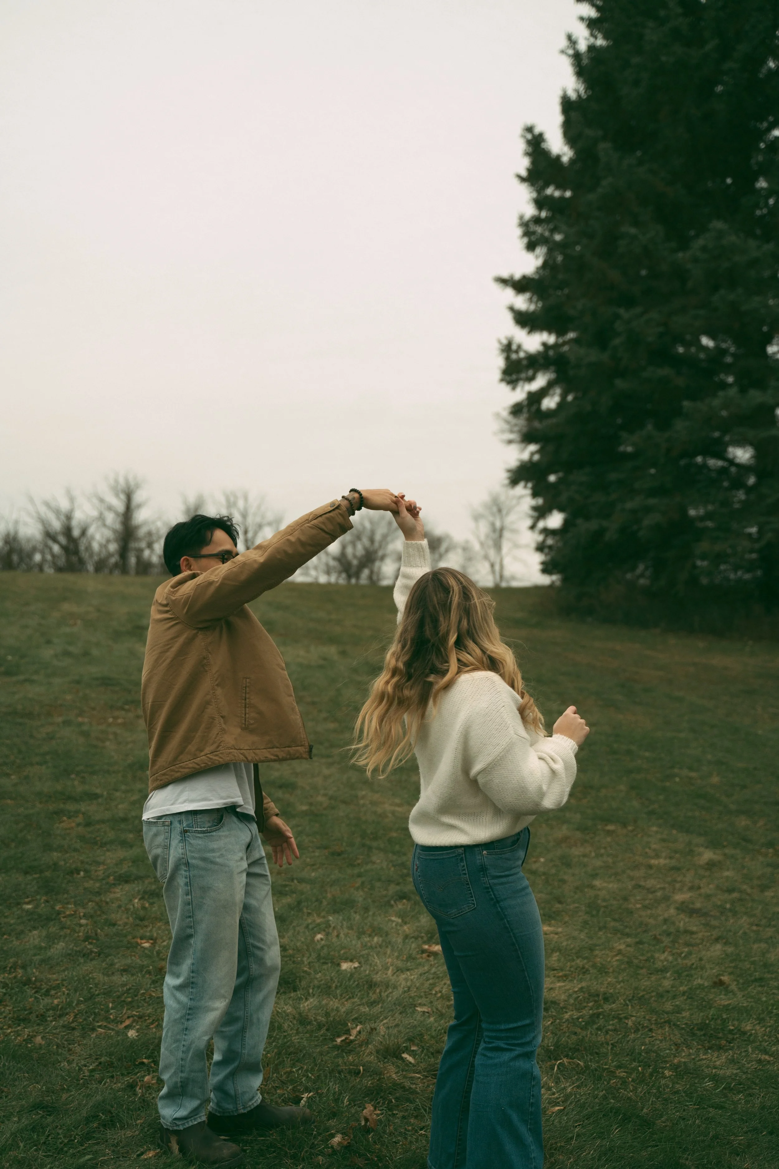 A man and a woman dancing outdoors on a grassy hill, with trees in the background, on an overcast day.