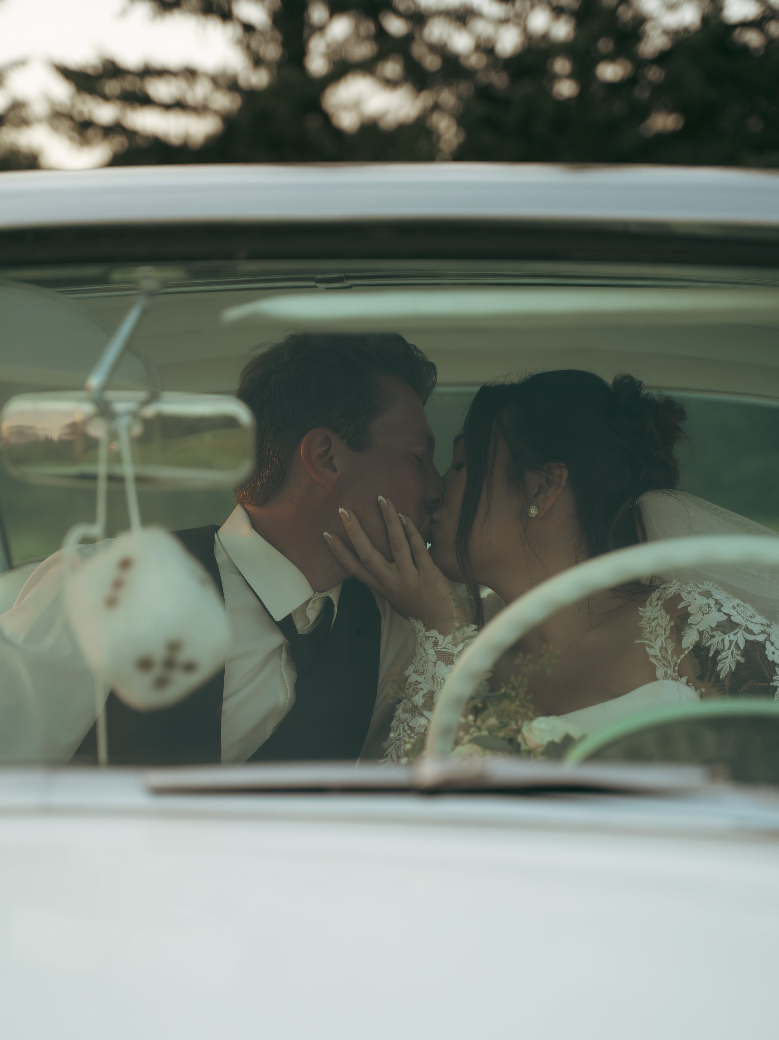 A couple kissing inside a vintage car, possibly on their wedding day, with trees visible through the windshield.
