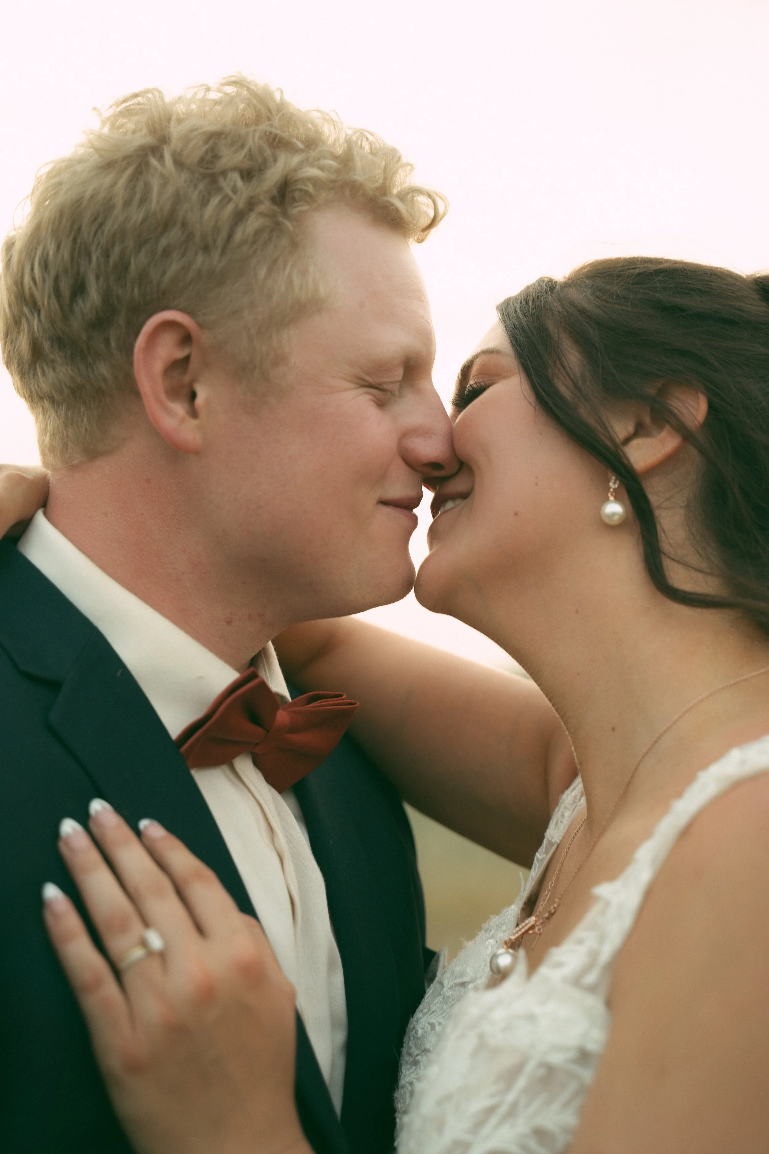 A close-up of a newlywed couple about to kiss, smiling with their eyes closed. The groom has curly blonde hair, a bow tie, and a suit. The bride has dark hair, pearl earrings, and is wearing a lace wedding dress. The bride's hand with a wedding ring is resting on the groom's shoulder.