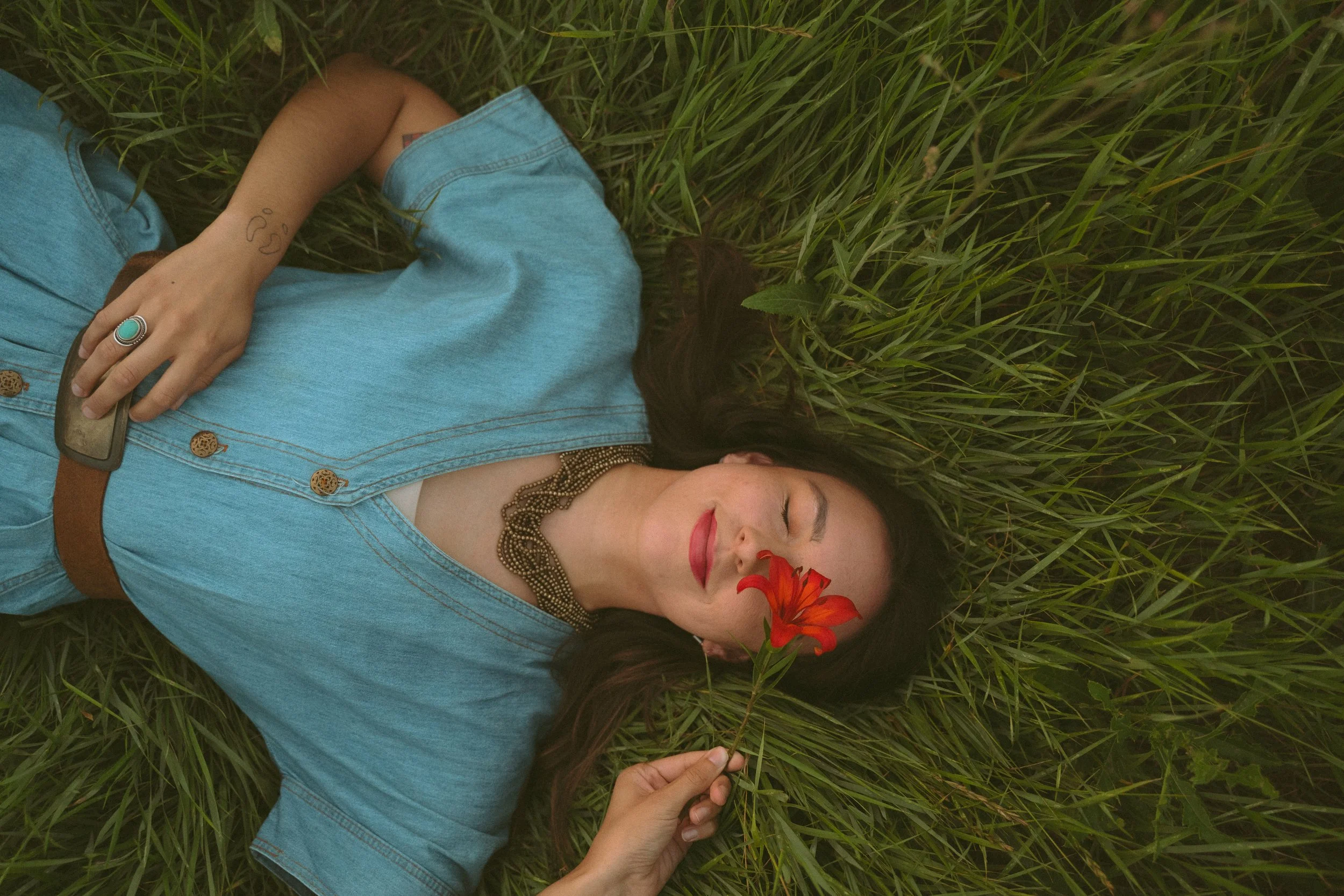 A woman lying in grass with her eyes closed, holding a red flower near her face, wearing a denim shirt, necklace, and ring.
