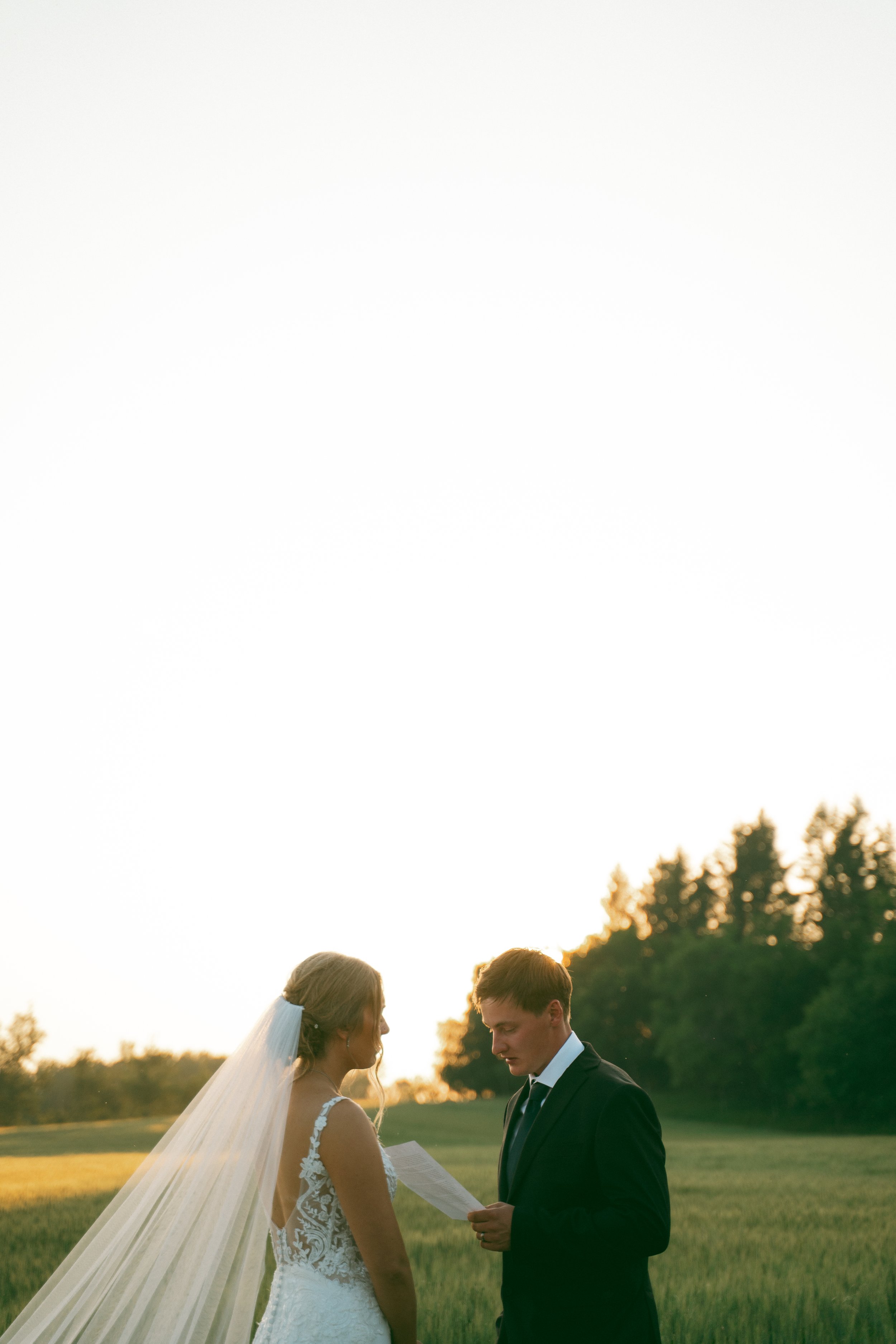 A bride and groom standing in a grassy field at sunset, with the bride holding a piece of paper and the groom reading it, in wedding attire.