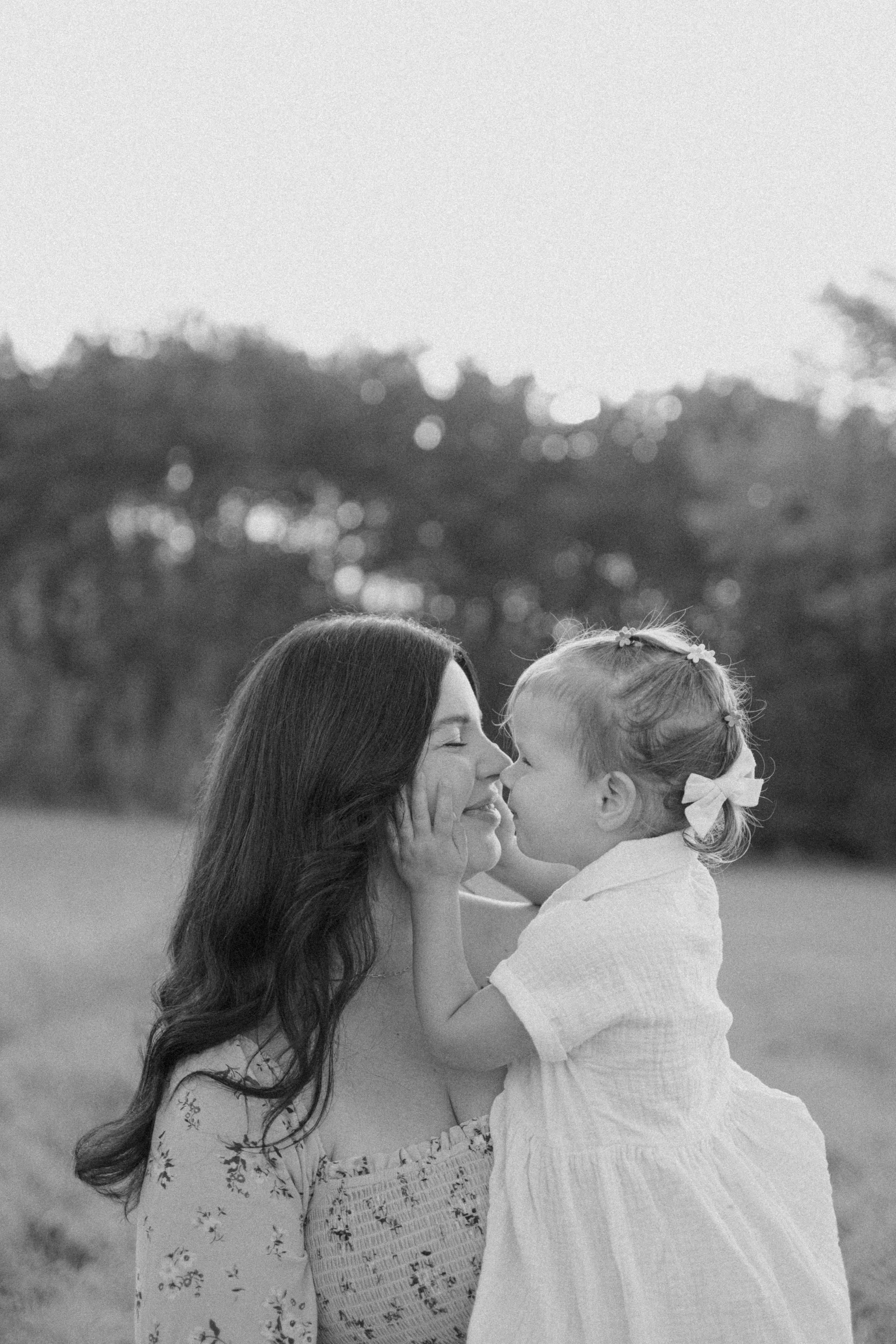 A woman and a young girl sharing a tender moment outdoors. The girl is touching the woman's face and they are close, with their noses almost touching. The background shows trees and a clear sky.