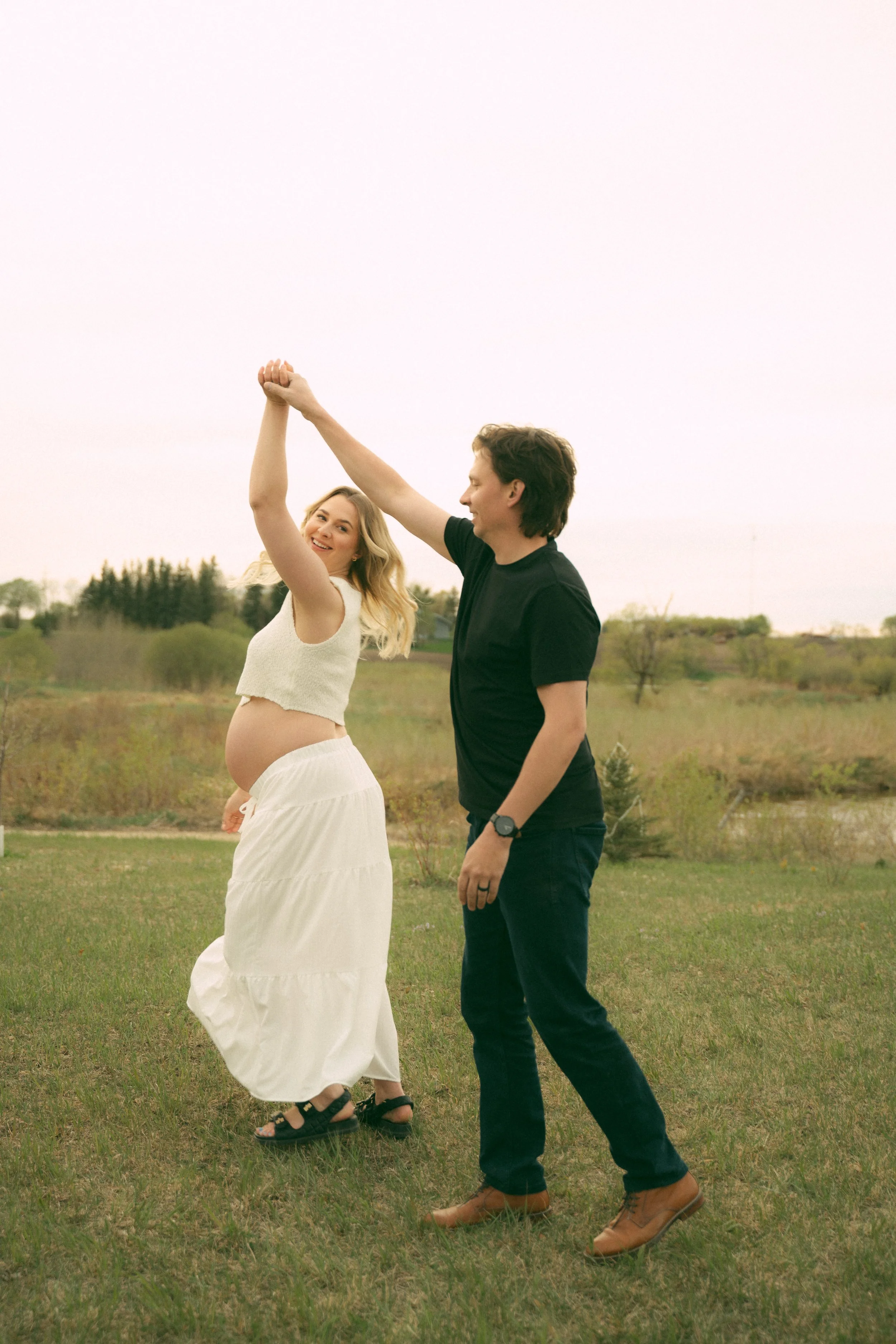 A pregnant woman in a white skirt and sleeveless top dancing with a man in a black t-shirt and jeans outdoors in a grassy field, holding hands and smiling.