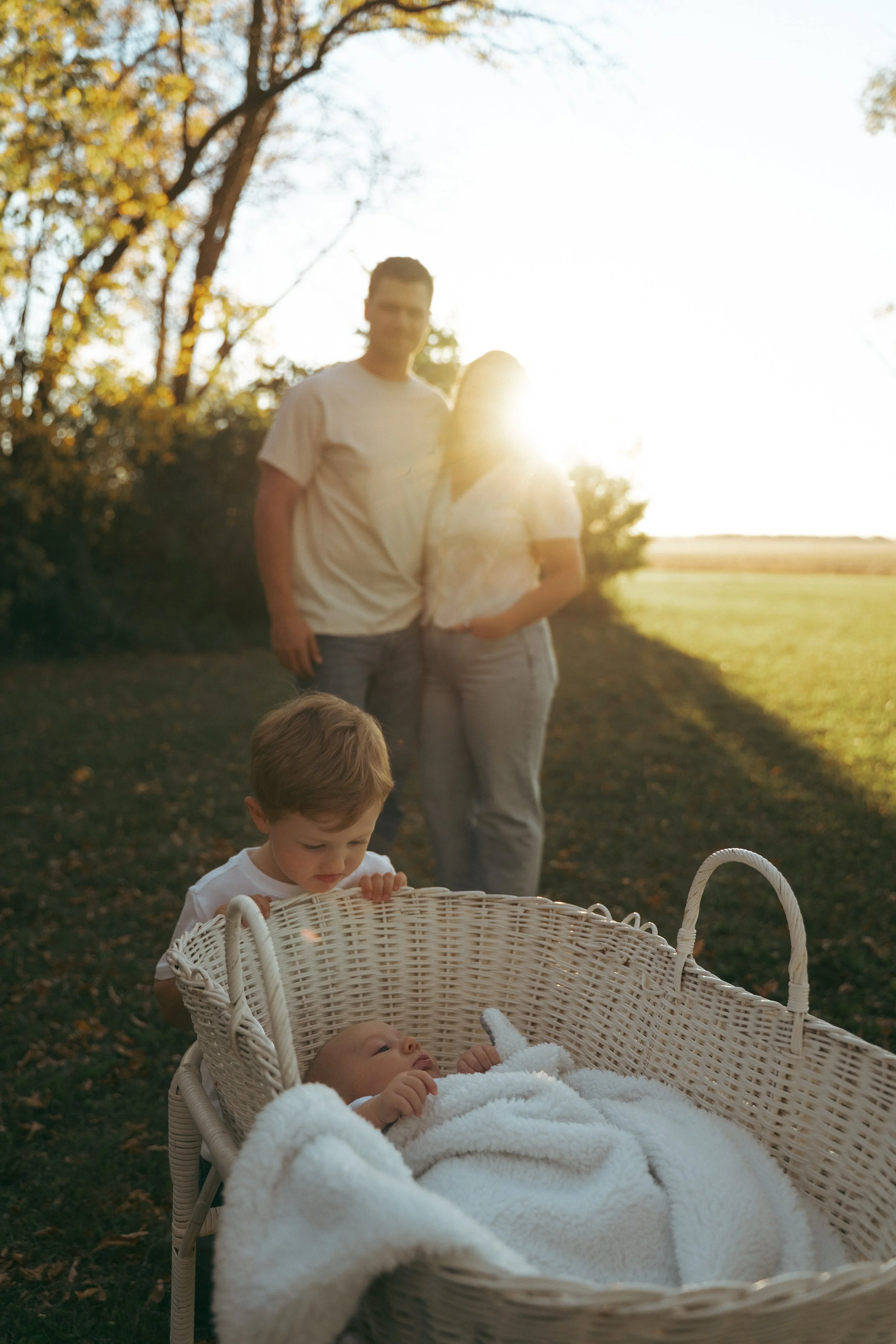 A young child and an older child or adult looking into a baby in a white blanket in a wicker bassinet outdoors at sunset.
