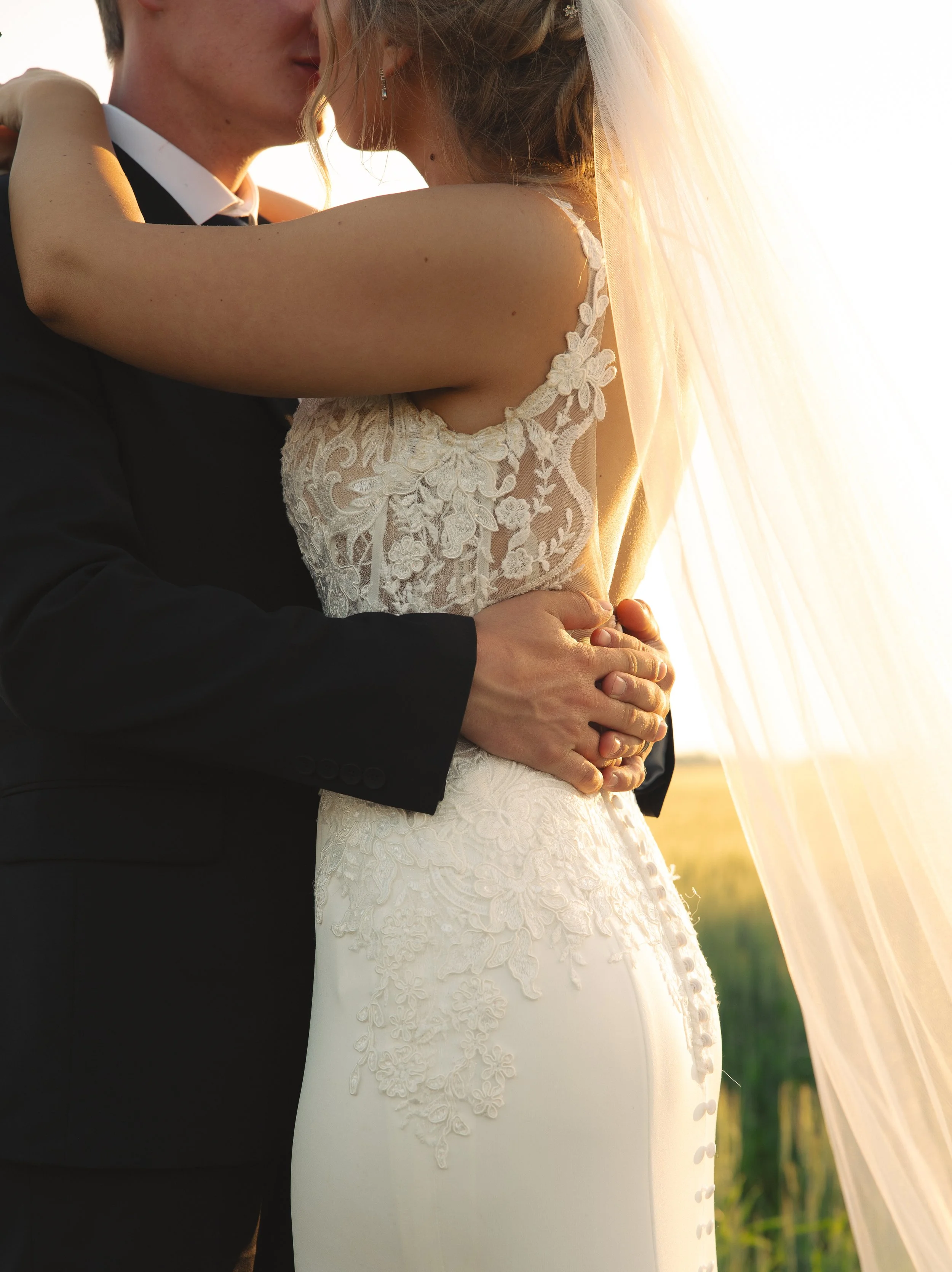 A bride and groom embracing outdoors during sunset, the bride in a white lace wedding dress with a veil, the groom in a black suit with a white shirt.