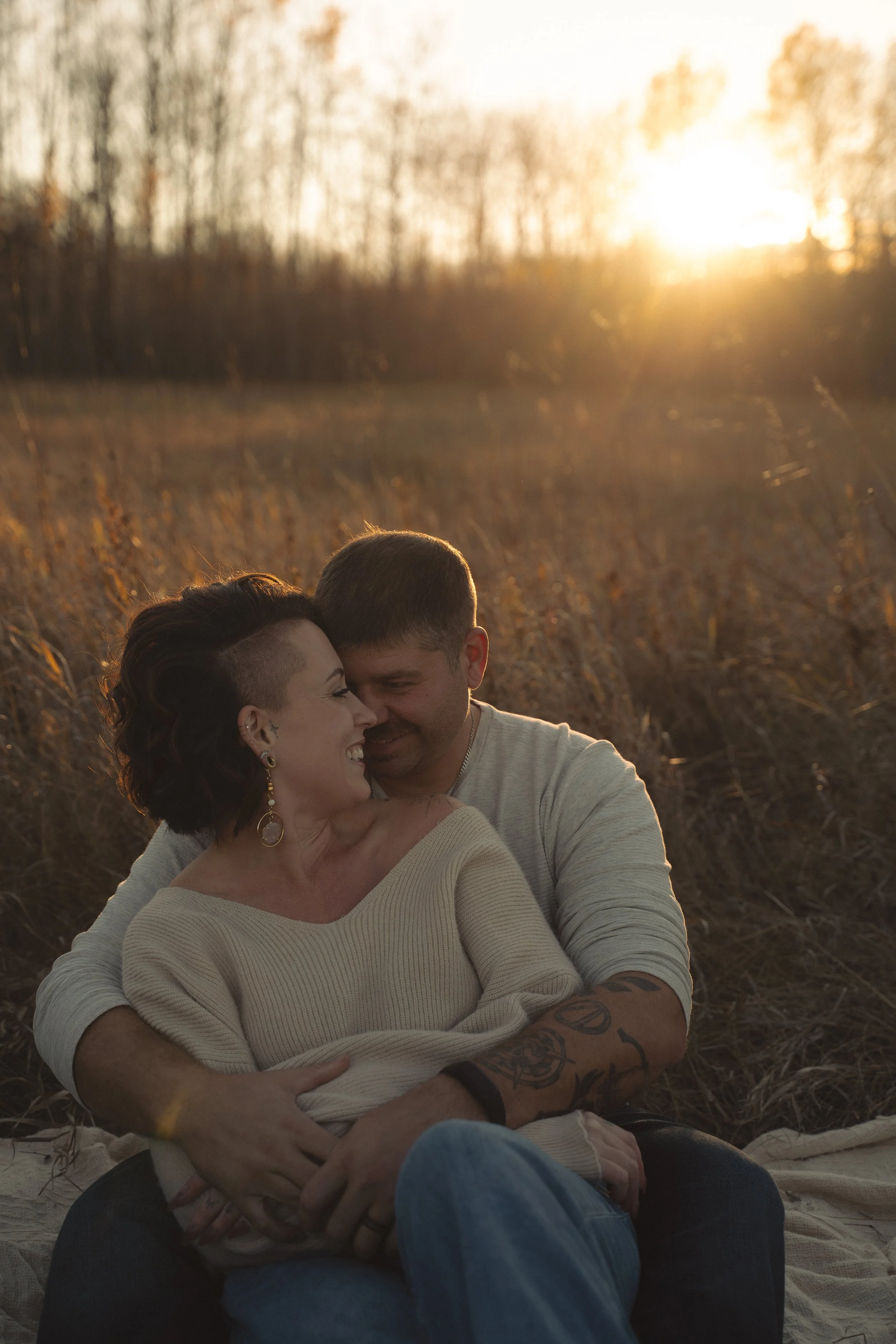 A couple sitting outdoors in a field during sunset, smiling and embracing each other.