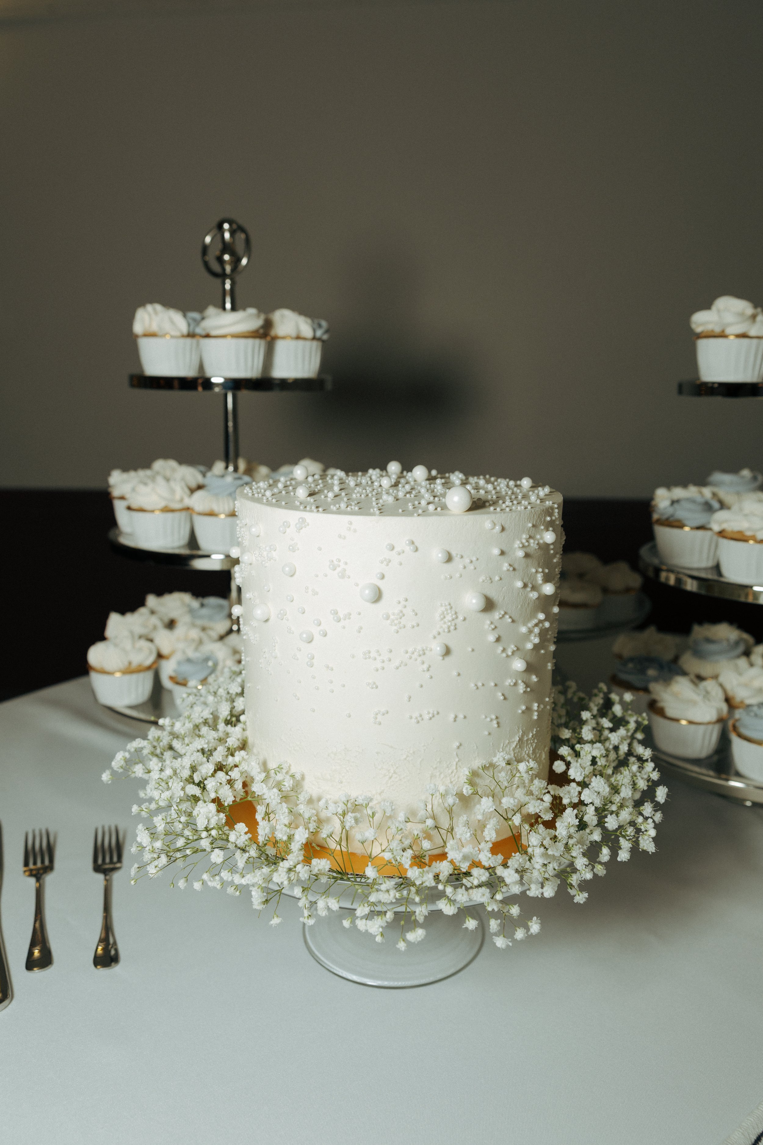 A white wedding cake decorated with small white pearls and tiny white flowers, surrounded by white baby's breath flowers at the base, with cupcake tiers in the background.