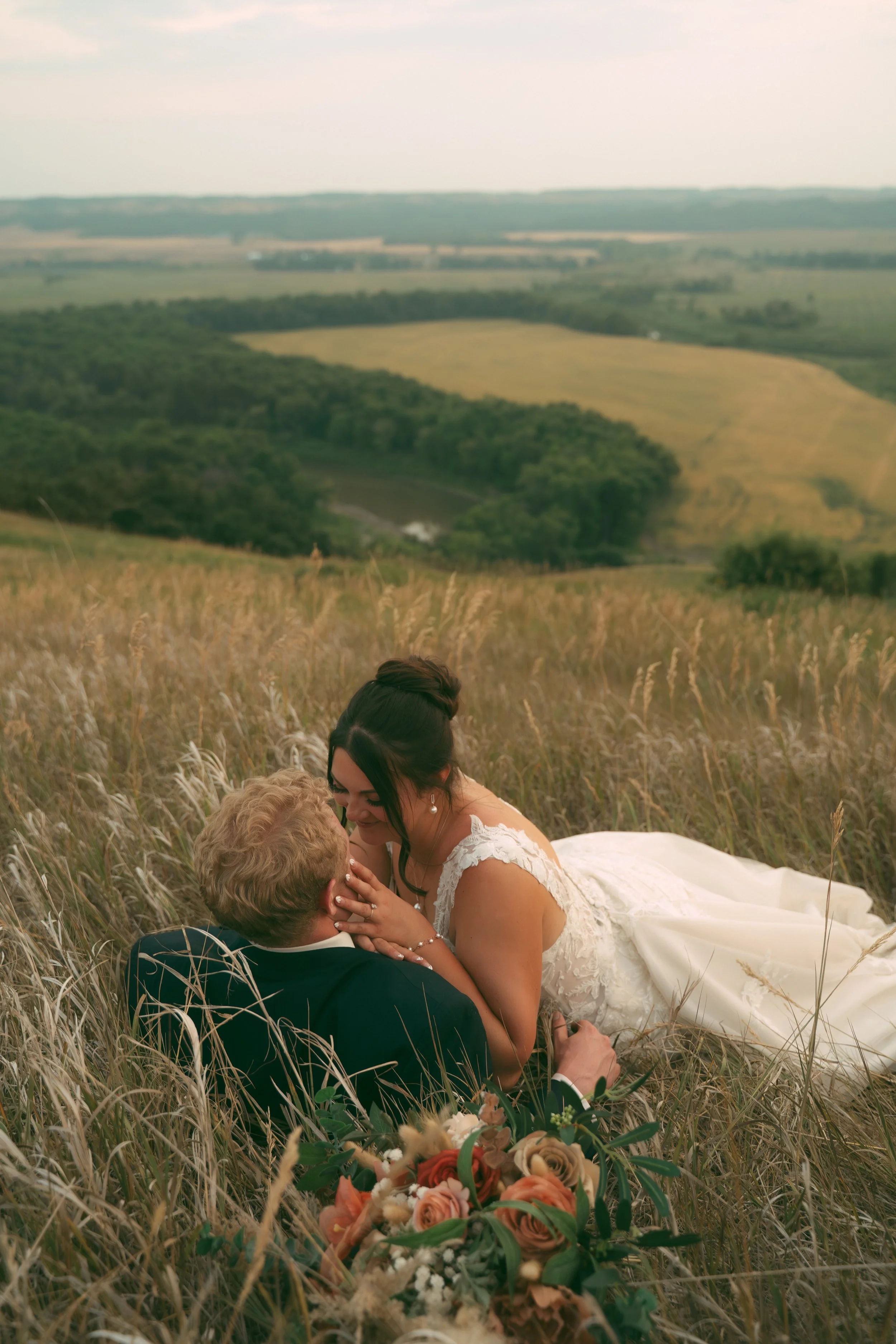 Bride and groom sitting in a field with tall grass, sharing a romantic moment with the bride touching the groom's face, and a bouquet of flowers in the foreground, with rolling hills and a cloudy sky in the background.