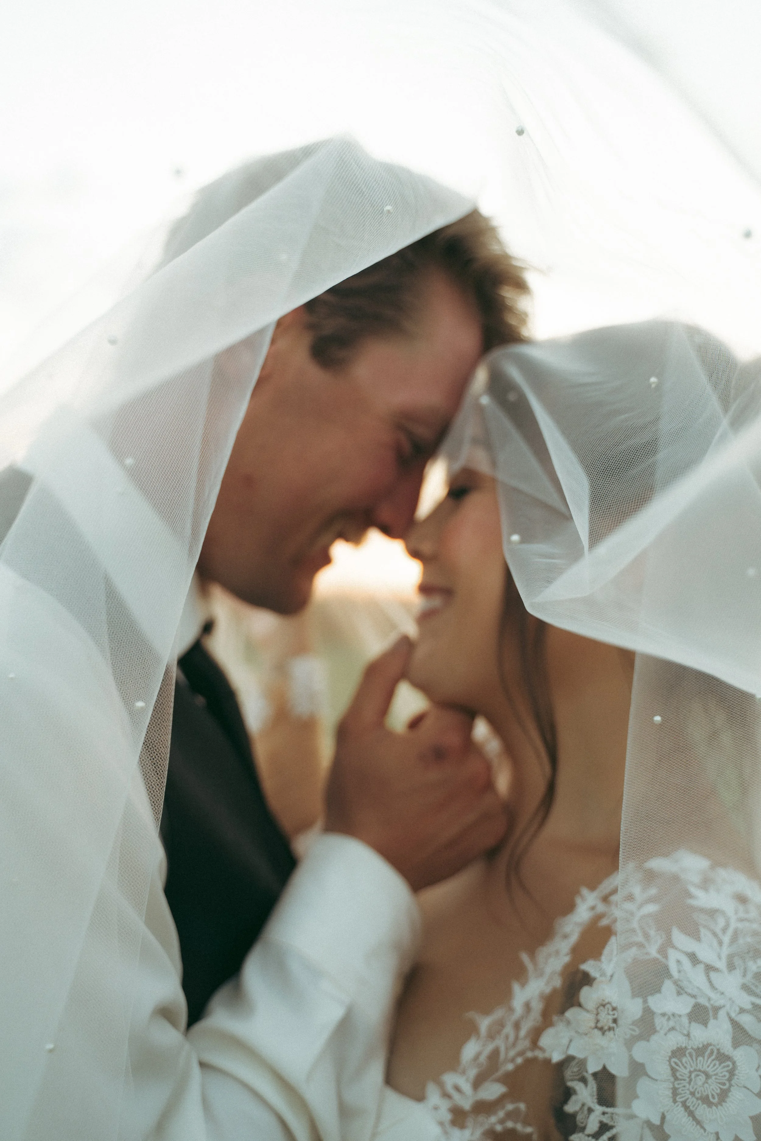 A couple on their wedding day, close-up, under a veil, smiling and touching foreheads, with soft natural lighting.