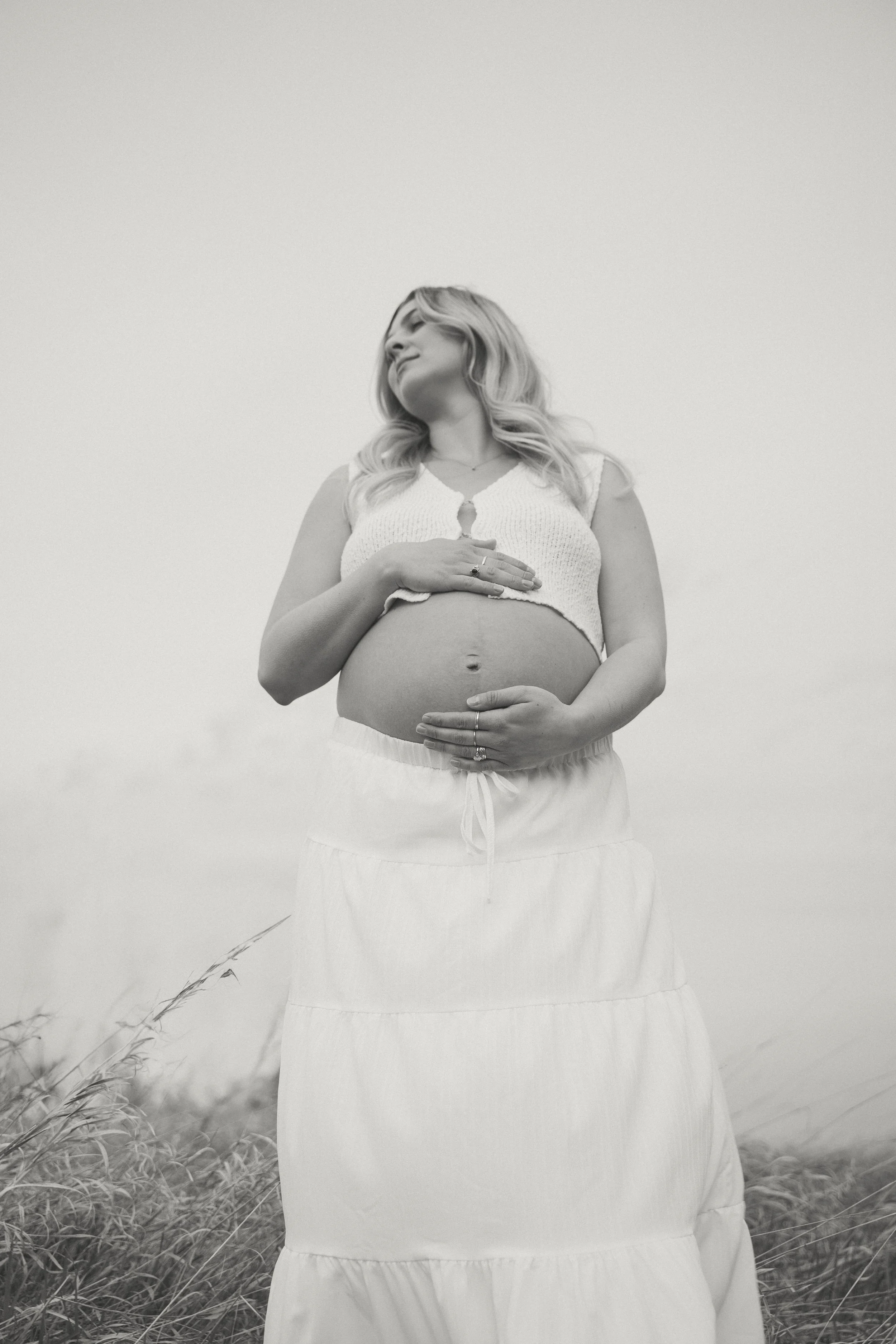 Black and white photo of a pregnant woman standing outdoors, cradling her belly with one hand on top and another below, looking slightly upward with a serene expression.