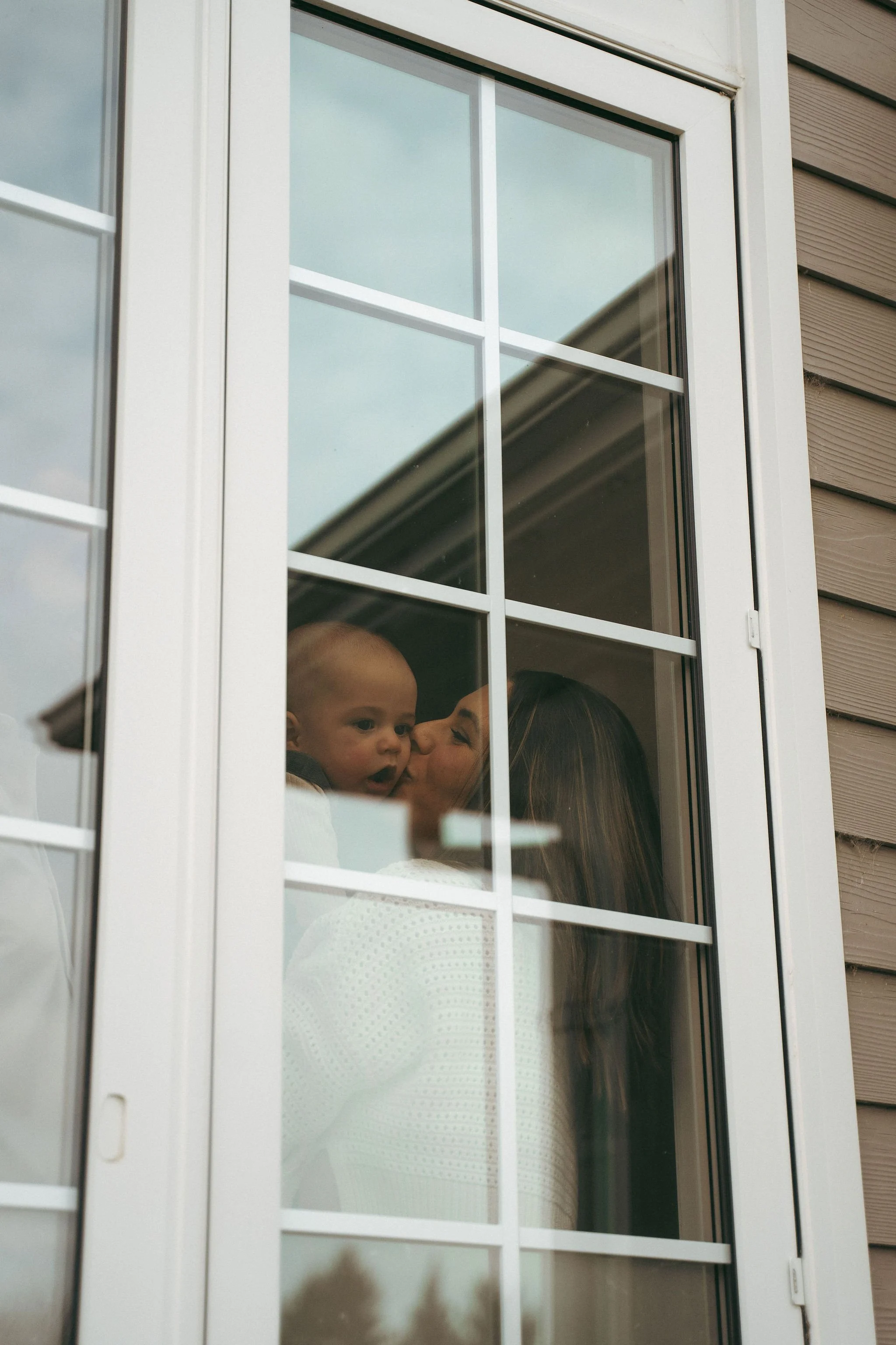 A woman holding a baby is seen through a window, with the woman giving a kiss to the baby's cheek.
