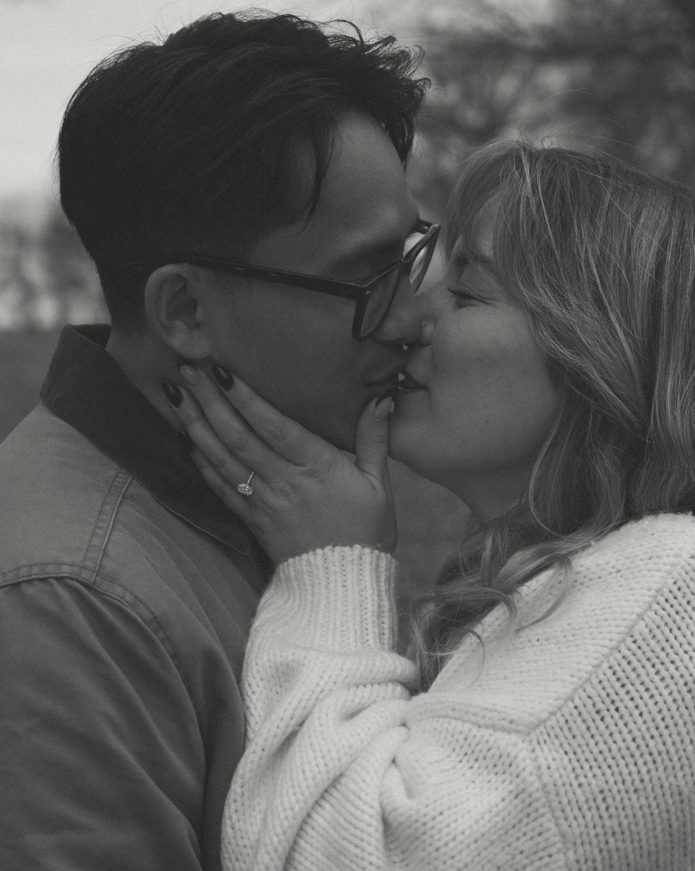 A black-and-white photo of a couple kissing; the woman gently holds the man's face with her hands, and they share a tender moment outdoors.