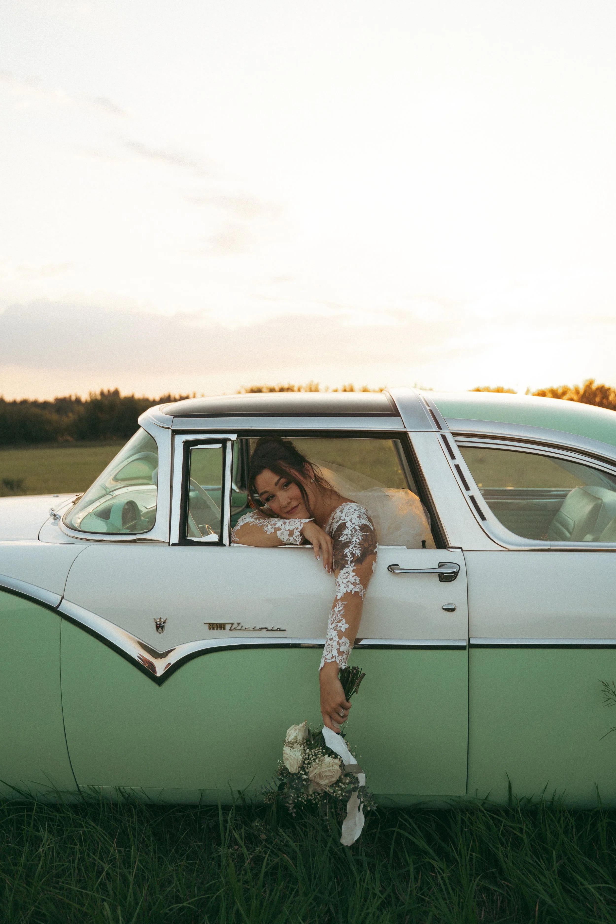 Young woman in wedding dress leaning out of vintage green and white car, holding bouquet, with countryside background at sunset.