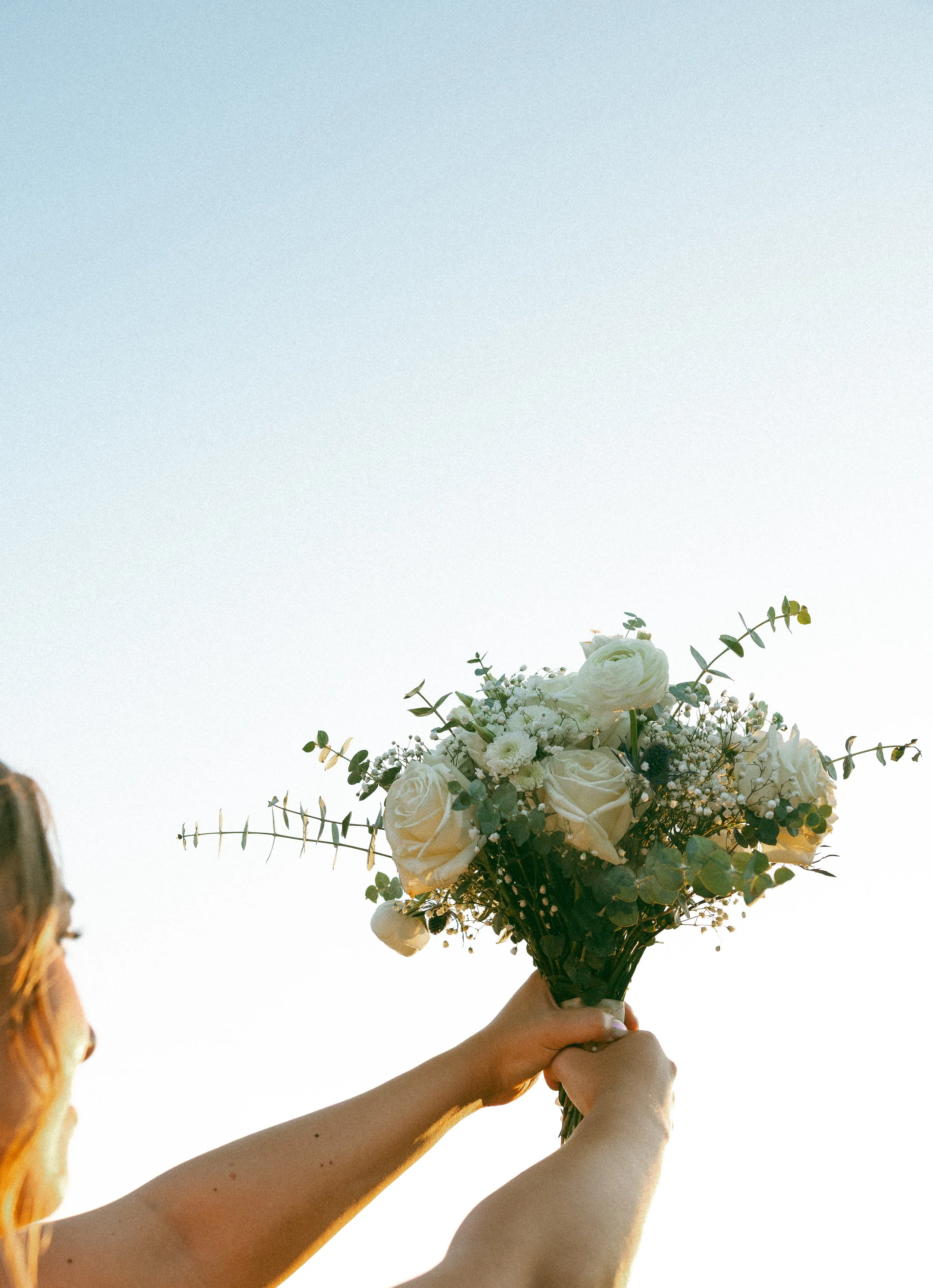 Person holding a bouquet of white roses and greenery against a clear sky.
