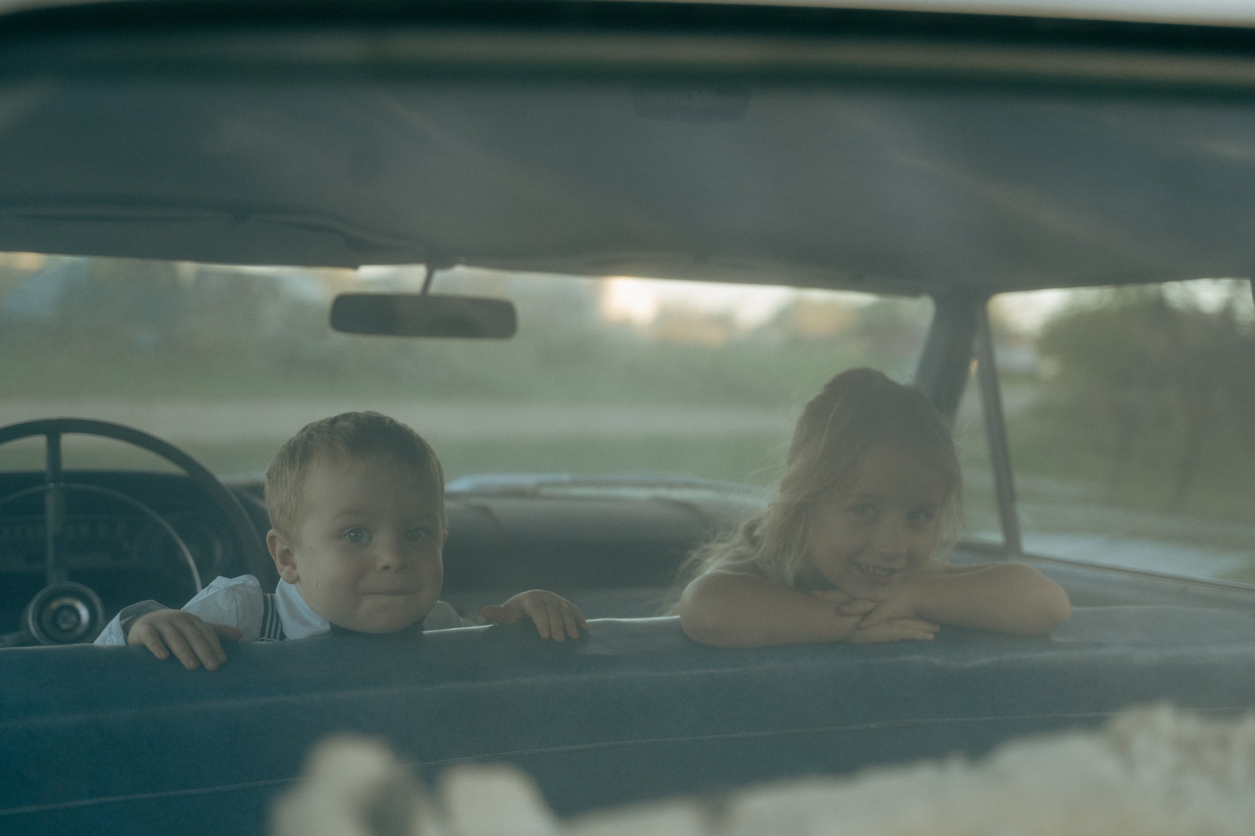 Two children leaning on the back of a car seat, smiling and looking outside the vehicle, with a blurred outdoor background.