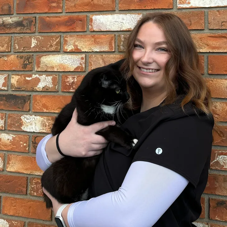A woman smiling and holding a black and white cat in front of a brick wall.