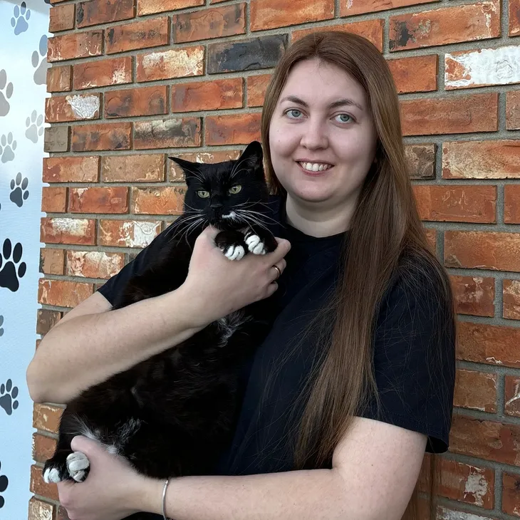 A woman with long brown hair holding a black and white cat with green eyes in front of a brick wall.