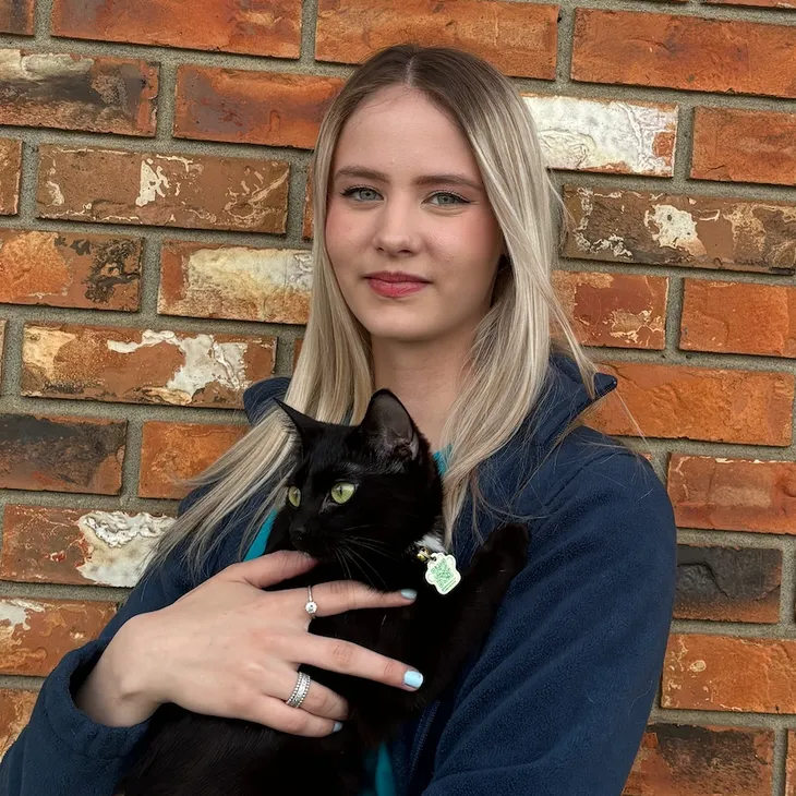 A young woman with long blonde hair holds a black cat with bright green eyes against a brick wall background.