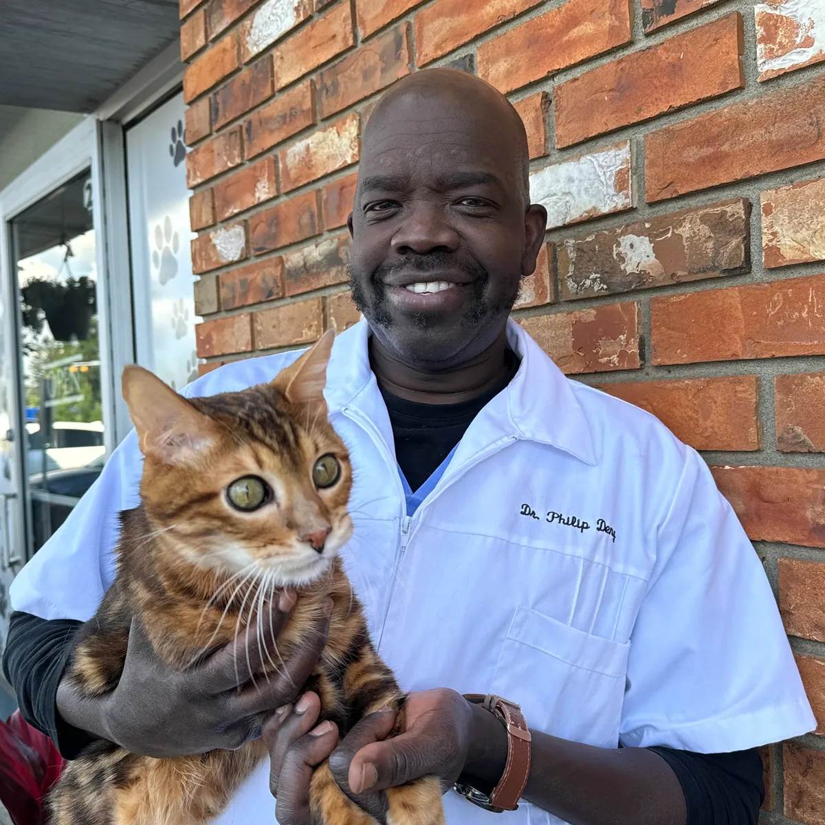 A man in a white coat holding a tabby cat outside a building with a brick wall background
