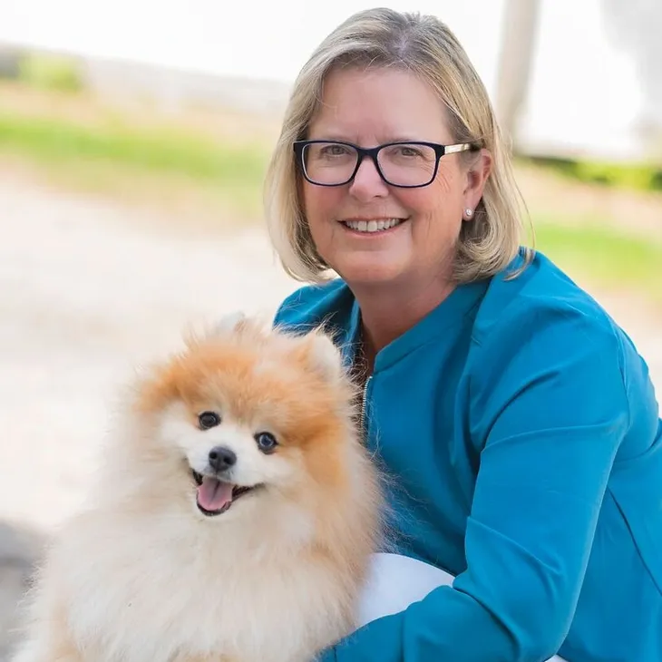 A woman with glasses and blond hair smiling and holding a fluffy Pomeranian dog outdoors.