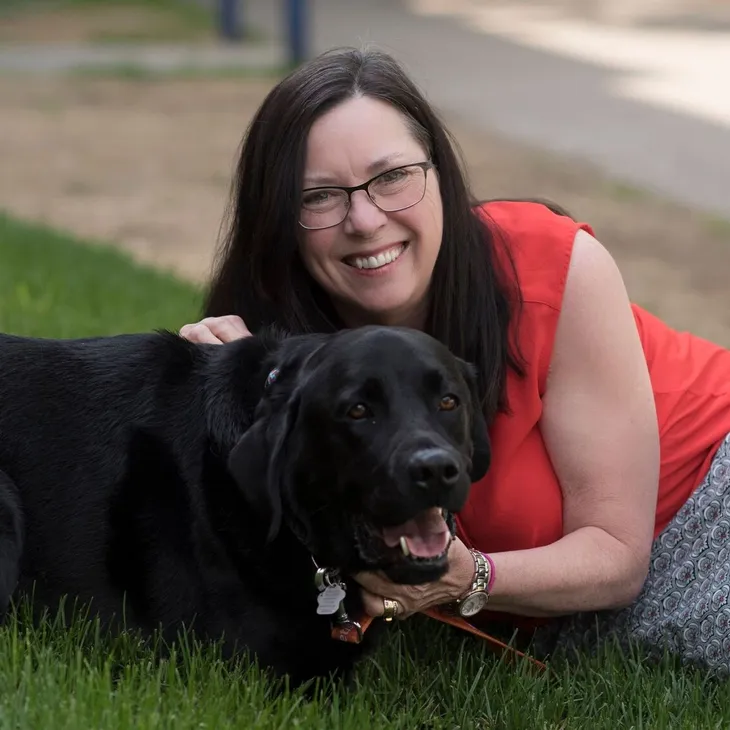 A woman with glasses and long dark hair lying on green grass, smiling while holding a black Labrador Retriever dog.