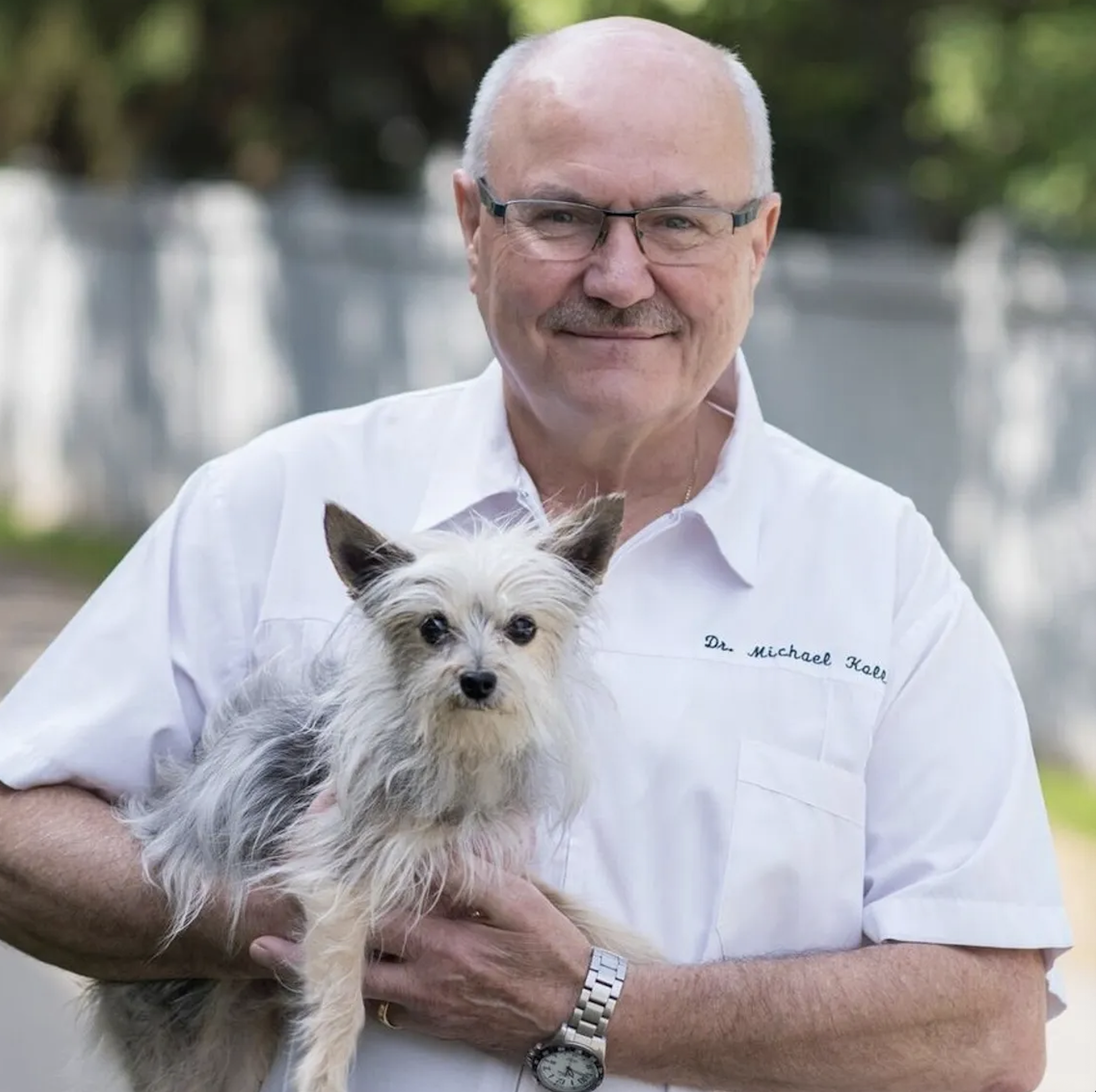 An elderly man with glasses and a white shirt holding a small, scruffy dog outdoors with a blurred background of trees and a fence.