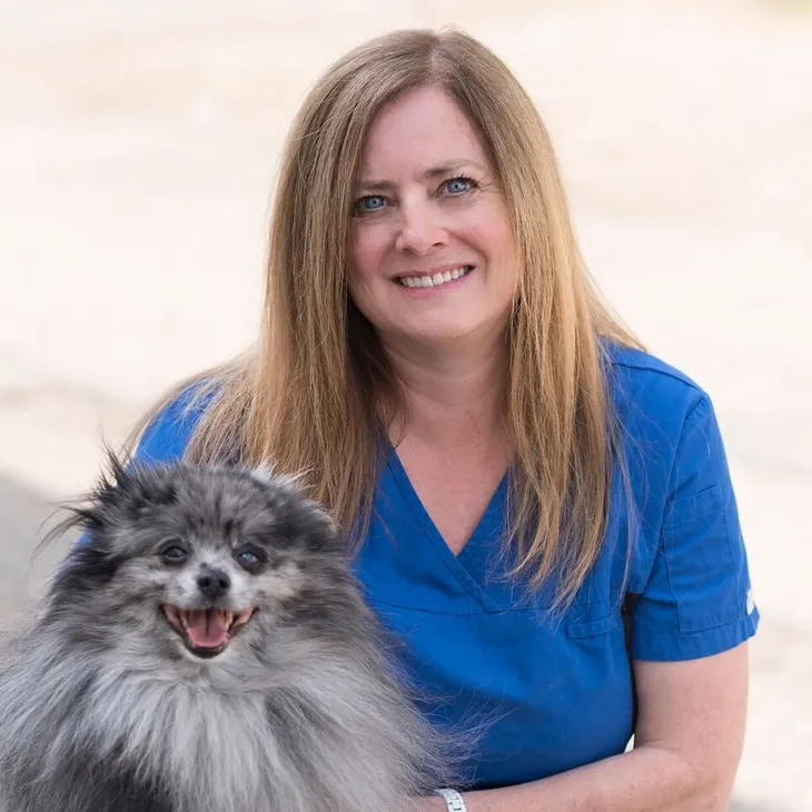 A woman with long reddish hair wearing a blue uniform, smiling, next to a fluffy gray dog with a happy expression.